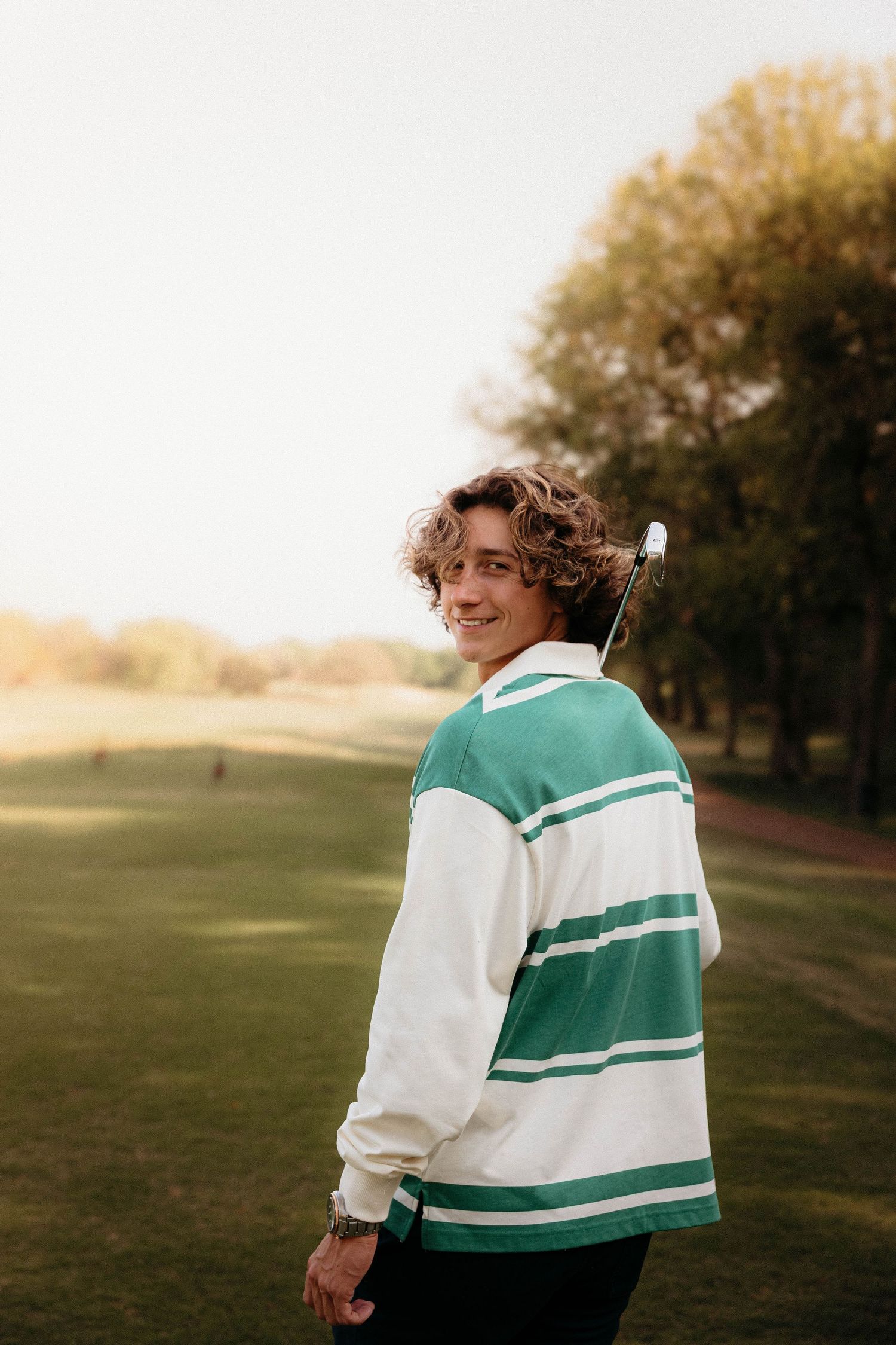 Young person in a green and white striped sweater smiling on a golf course at sunset.