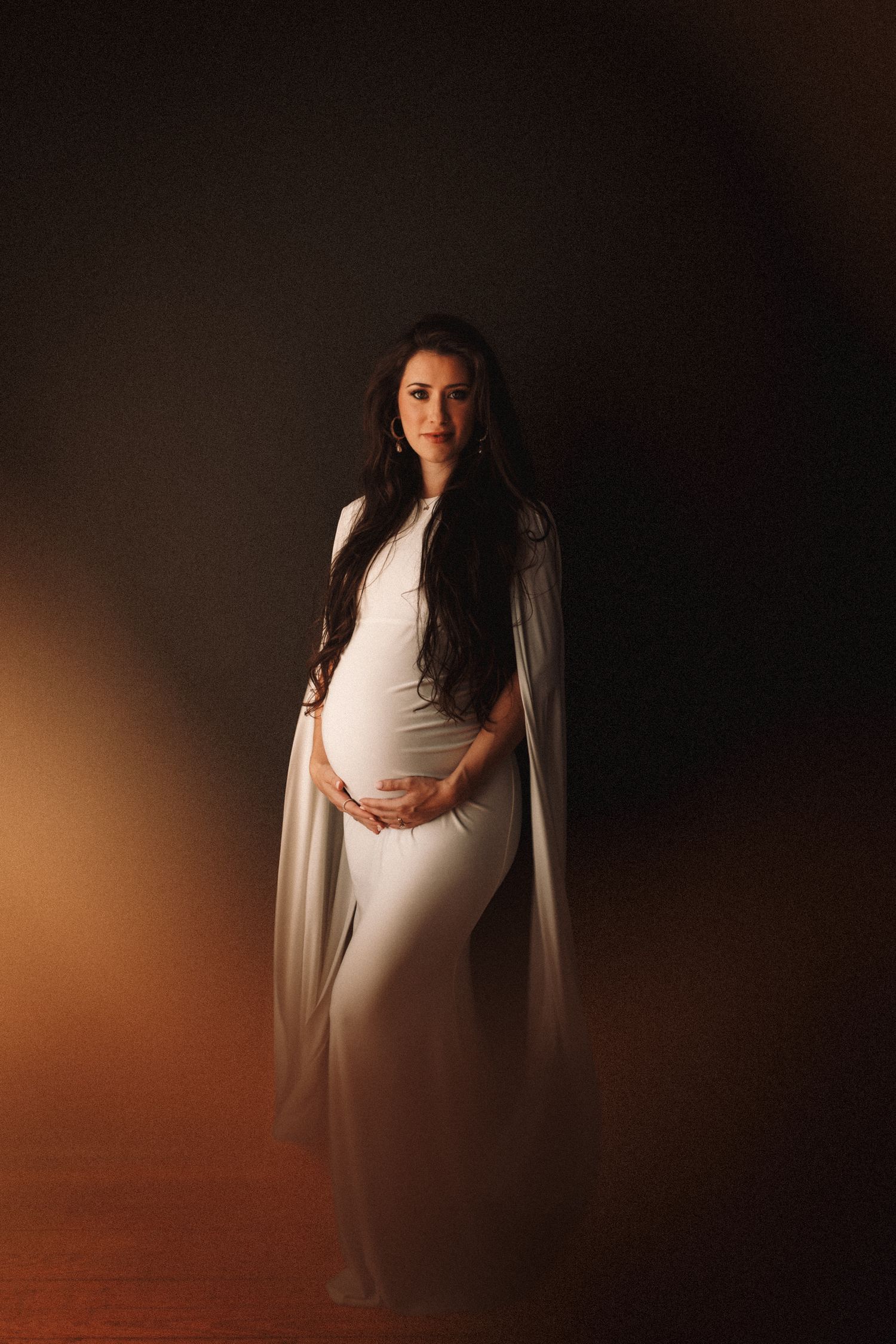 A dramatic maternity portrait featuring a flowing white gown and cape against a dark background with warm lighting.