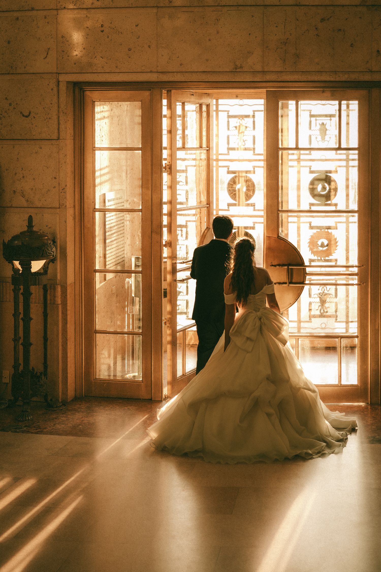 Bride and groom silhouetted against ornate golden stained glass doors in warm romantic wedding photo.