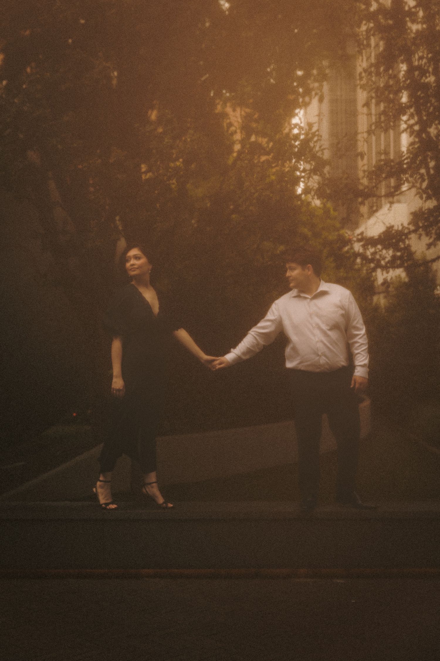 Couple holding hands in moody golden light outdoors, woman in black dress, man in white shirt, lush greenery behind.