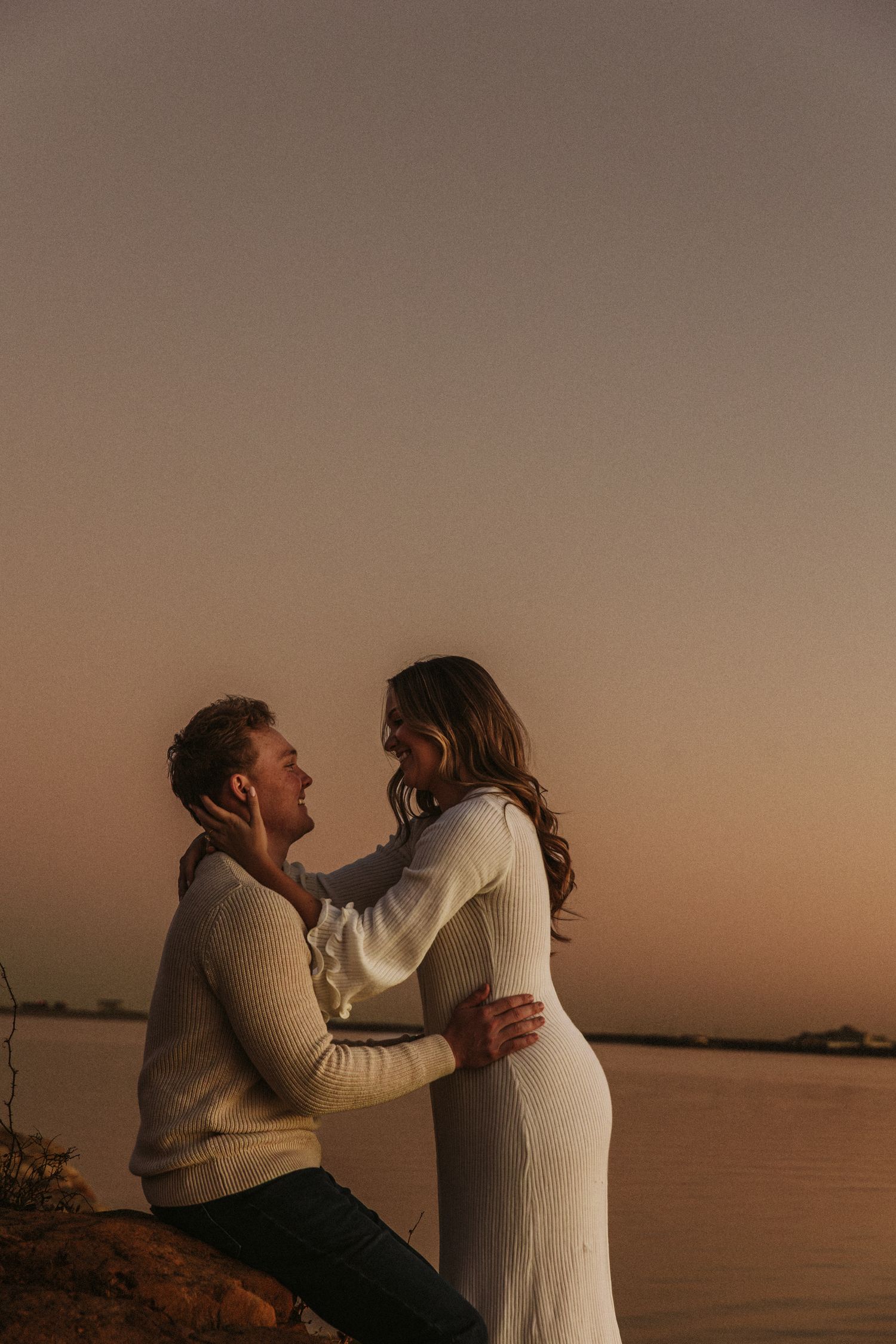 A romantic moment between a couple in white and grey outfits against a dusky waterfront backdrop.