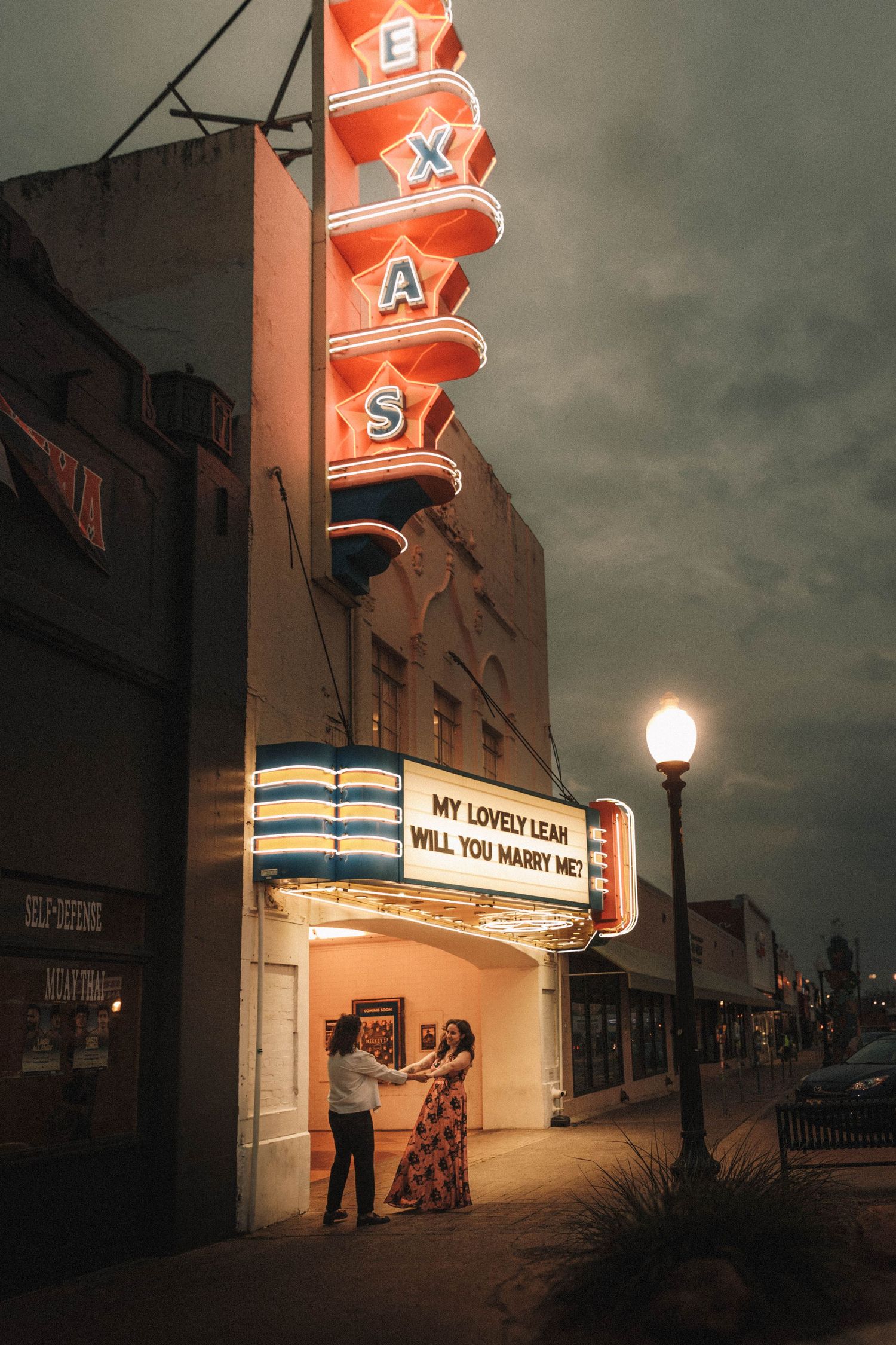 Couple dances outside the Texas Theatre at night under a glowing marriage proposal marquee sign.