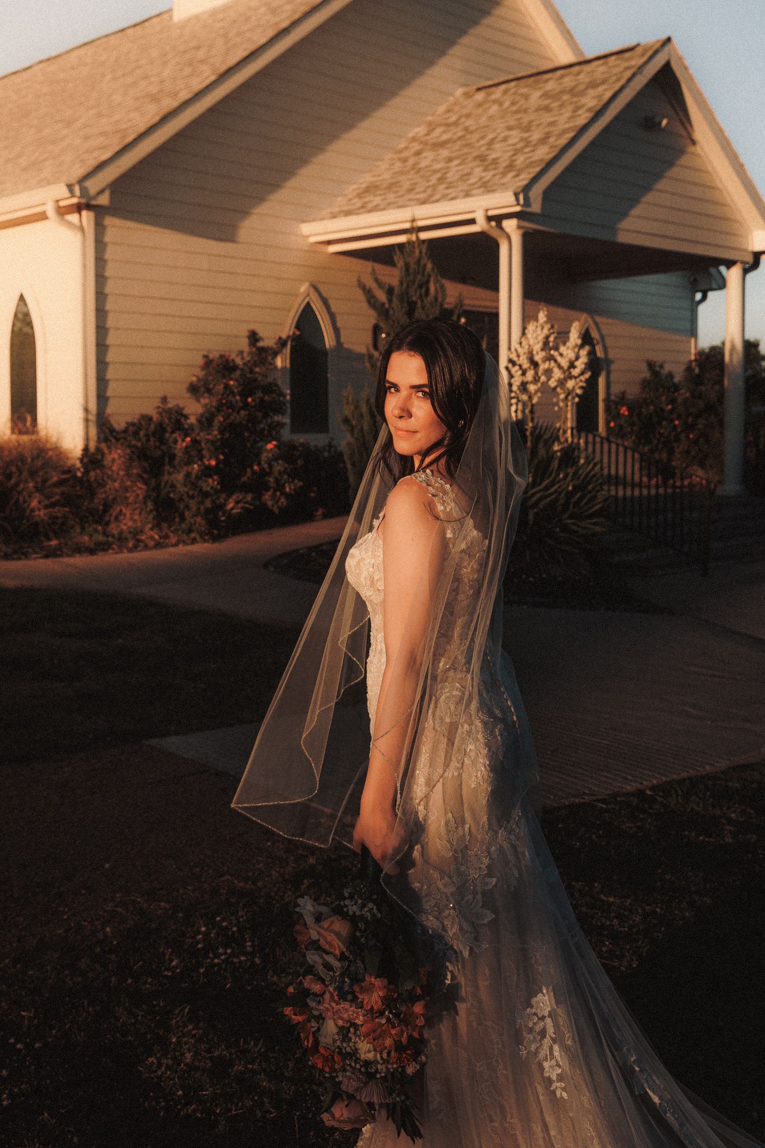 Bride in lace gown and flowing veil holding a dark bouquet at golden hour outside a white chapel.
