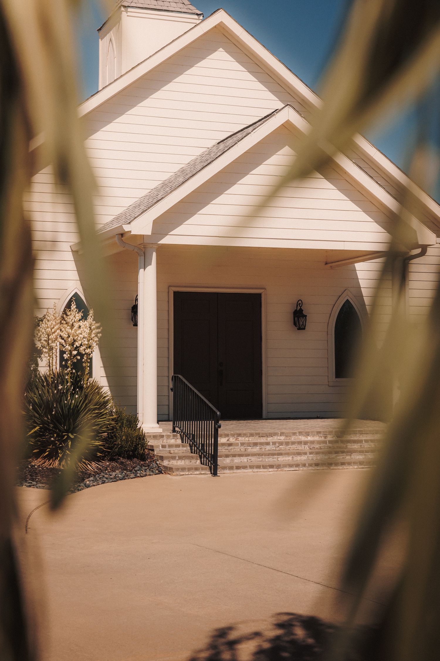 White chapel wedding venue with arched doorway, black railing steps, and manicured landscaping under a clear blue sky.