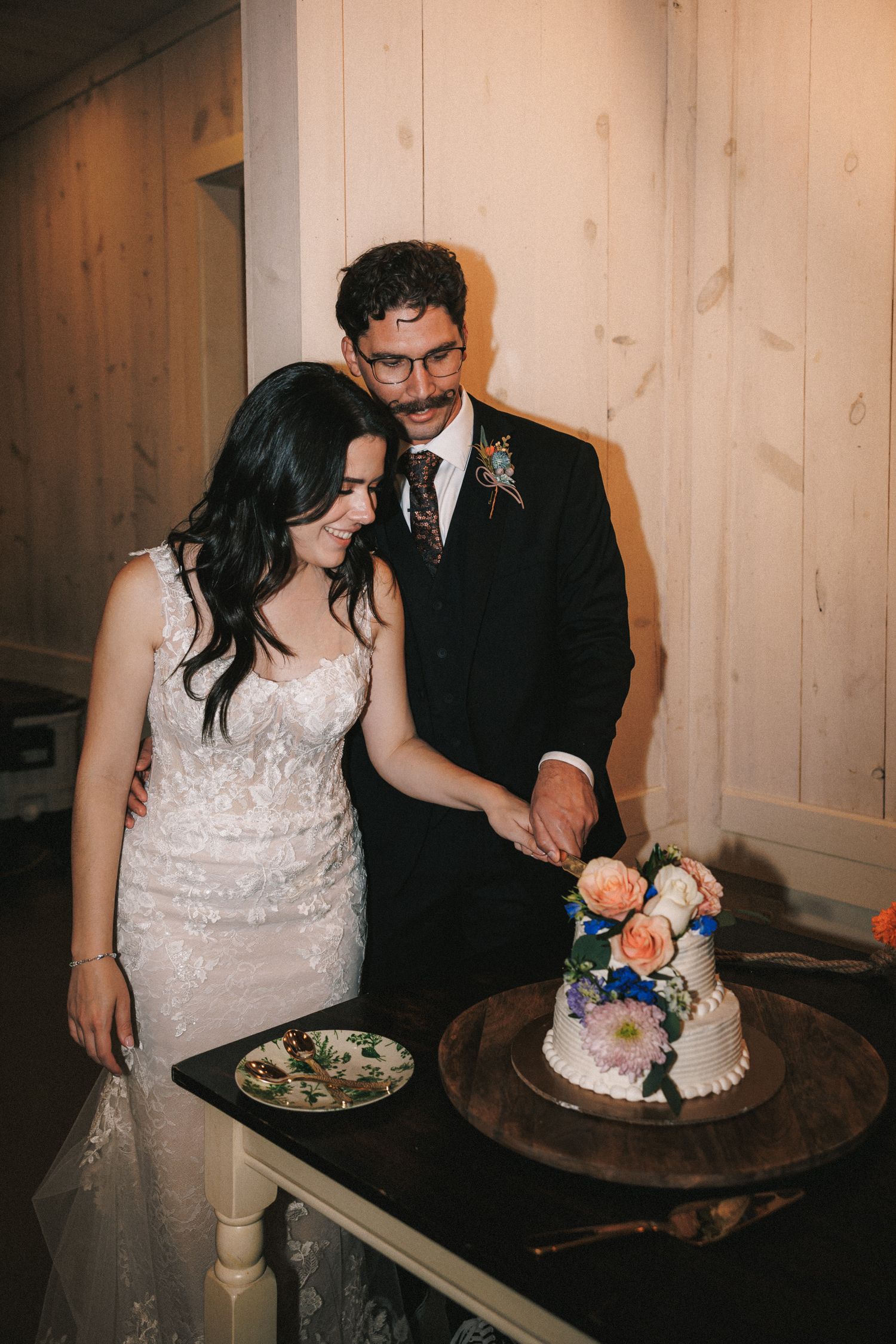 Bride and groom cut a floral-topped white wedding cake together inside a rustic wood-paneled venue.