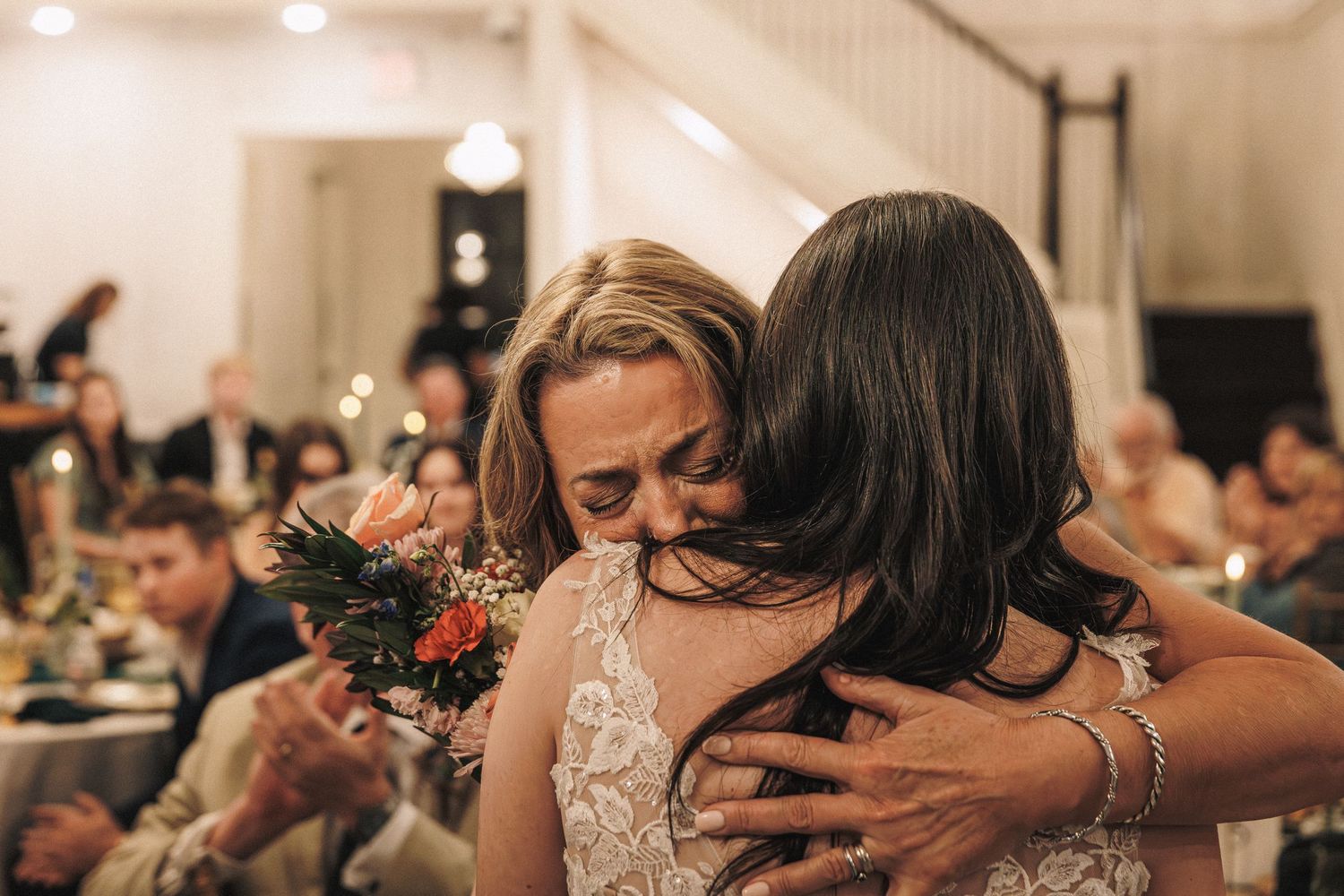 Emotional bride in lace gown sharing a heartfelt hug with a guest during the wedding reception.