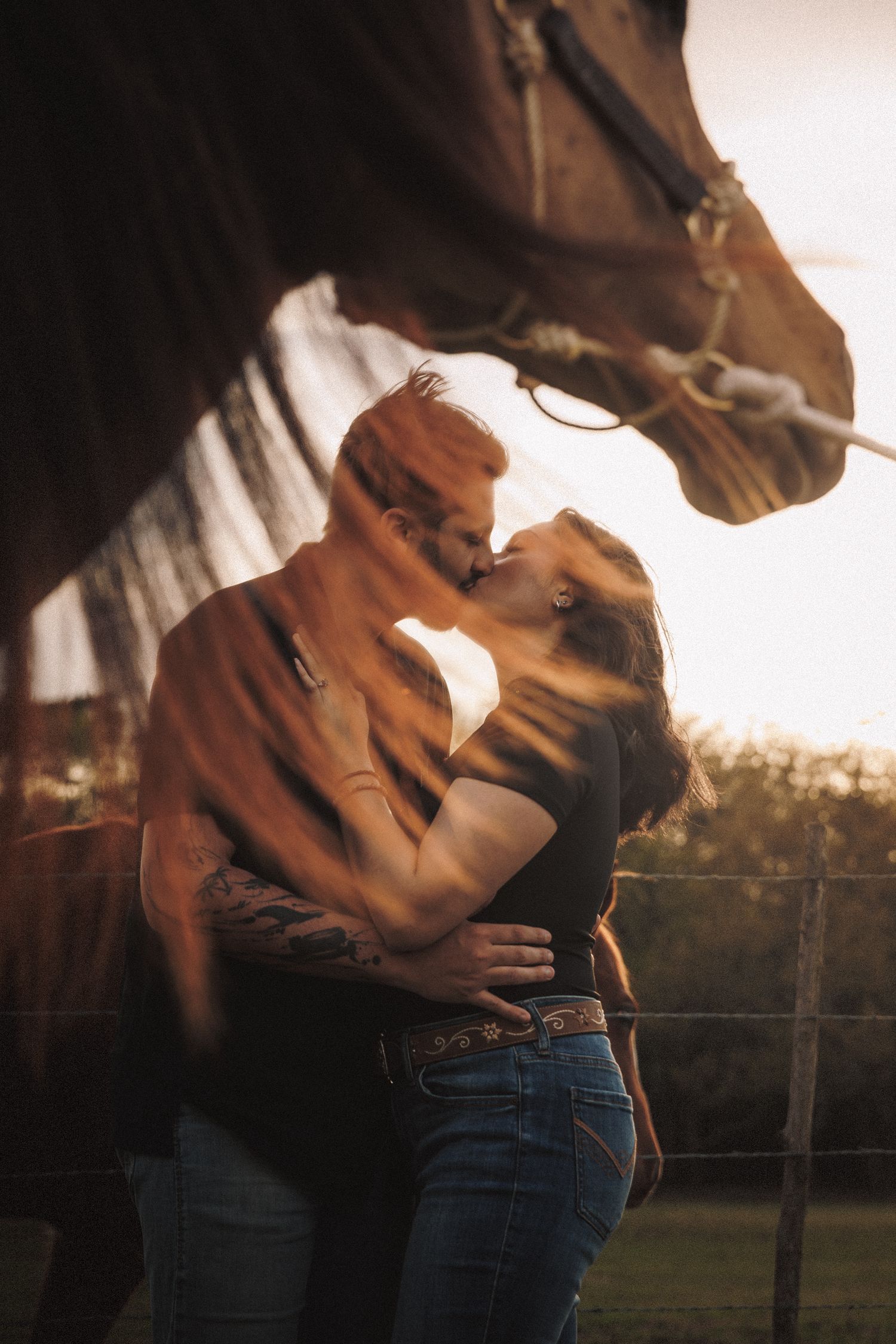 Couple sharing a passionate kiss framed by a horse at golden hour on a rustic farm.