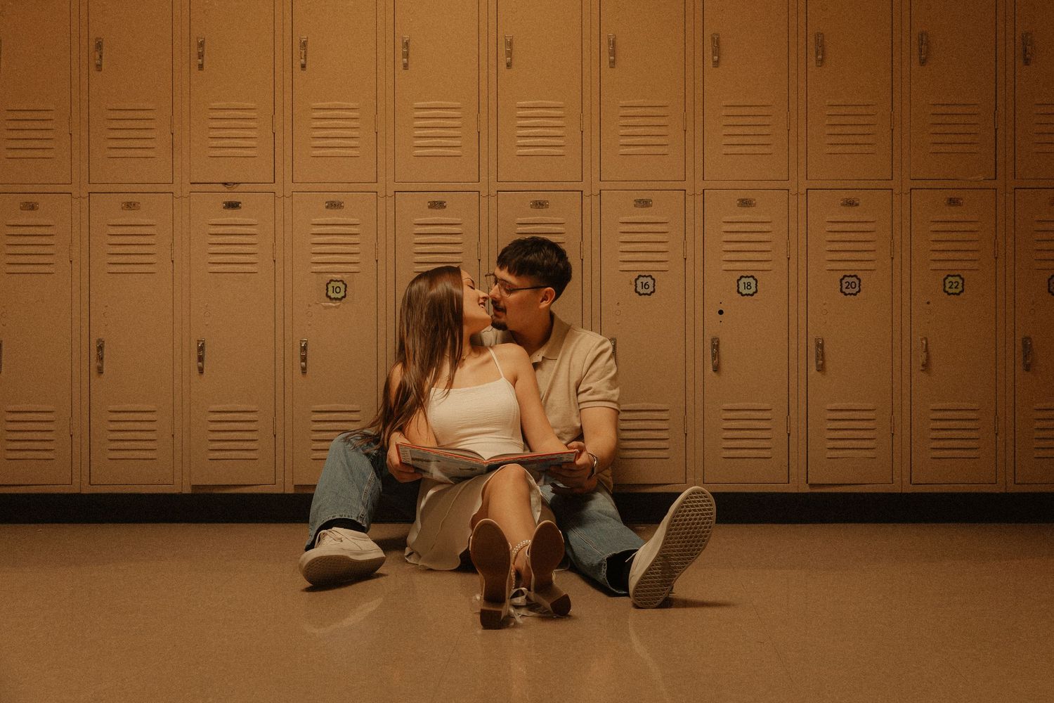 A couple sits on the floor sharing a kiss in front of beige school lockers in a warmly lit hallway.