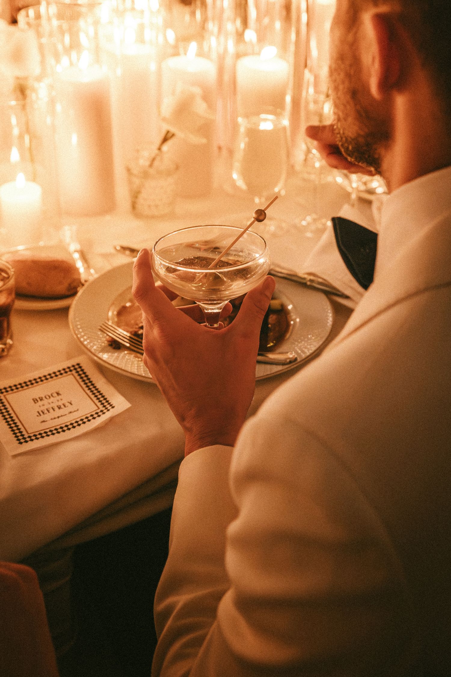 Groom in white tuxedo holding a martini at an elegant candlelit wedding reception dinner table.