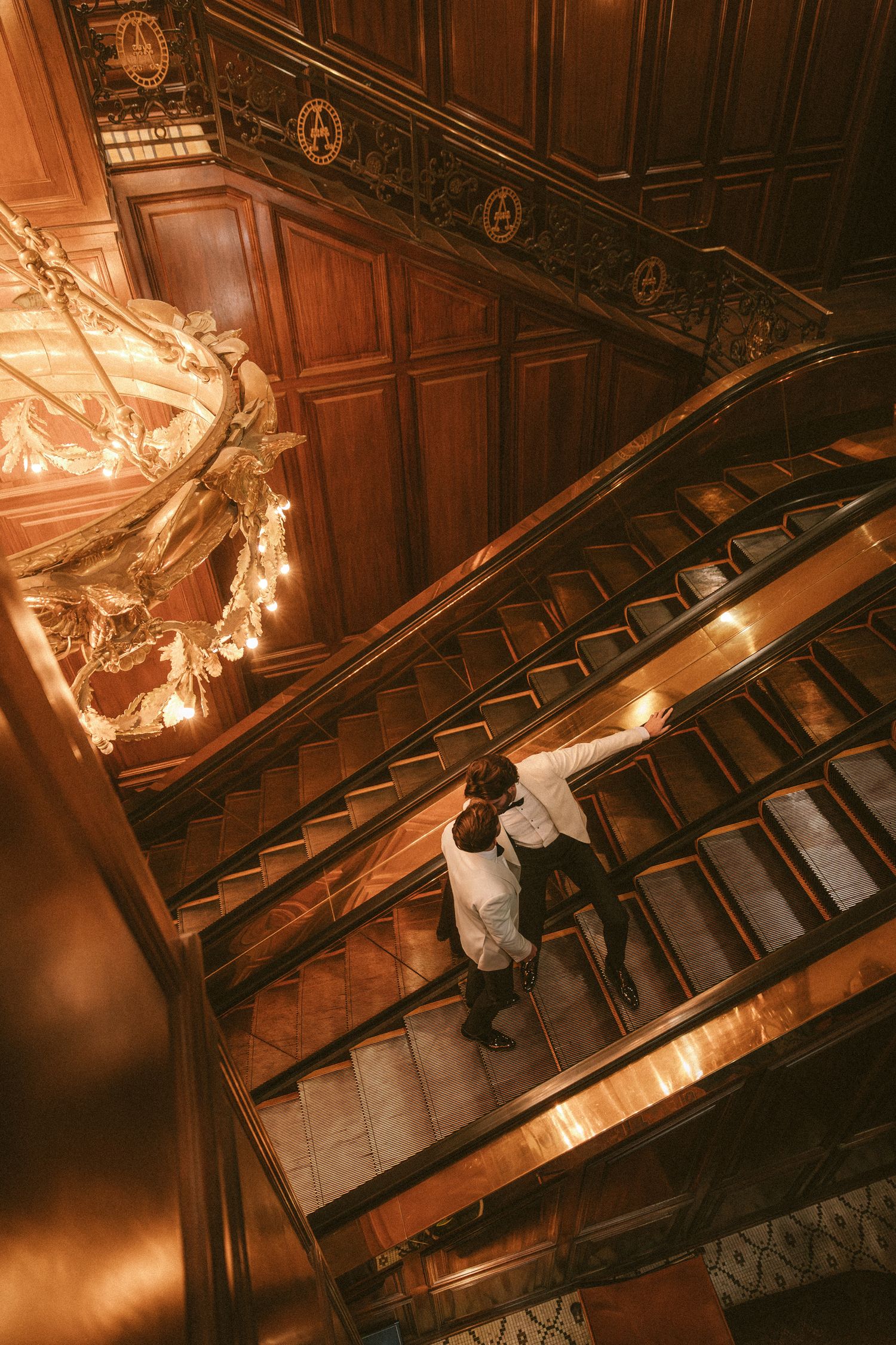 Overhead view of two grooms in white suits embracing on a grand ornate wooden staircase with chandelier.