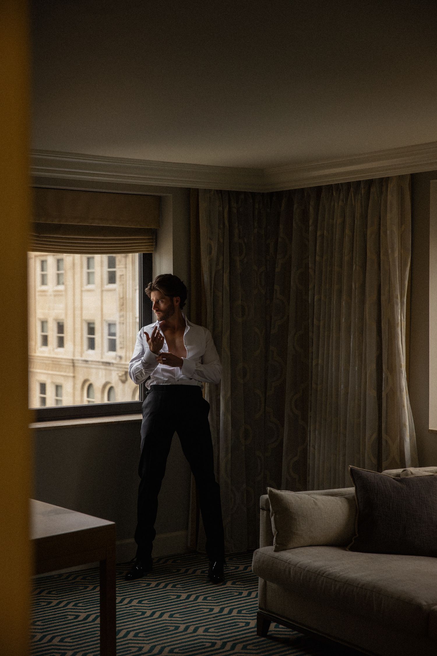 Groom buttoning white dress shirt in luxury hotel room with curtains and city view window.