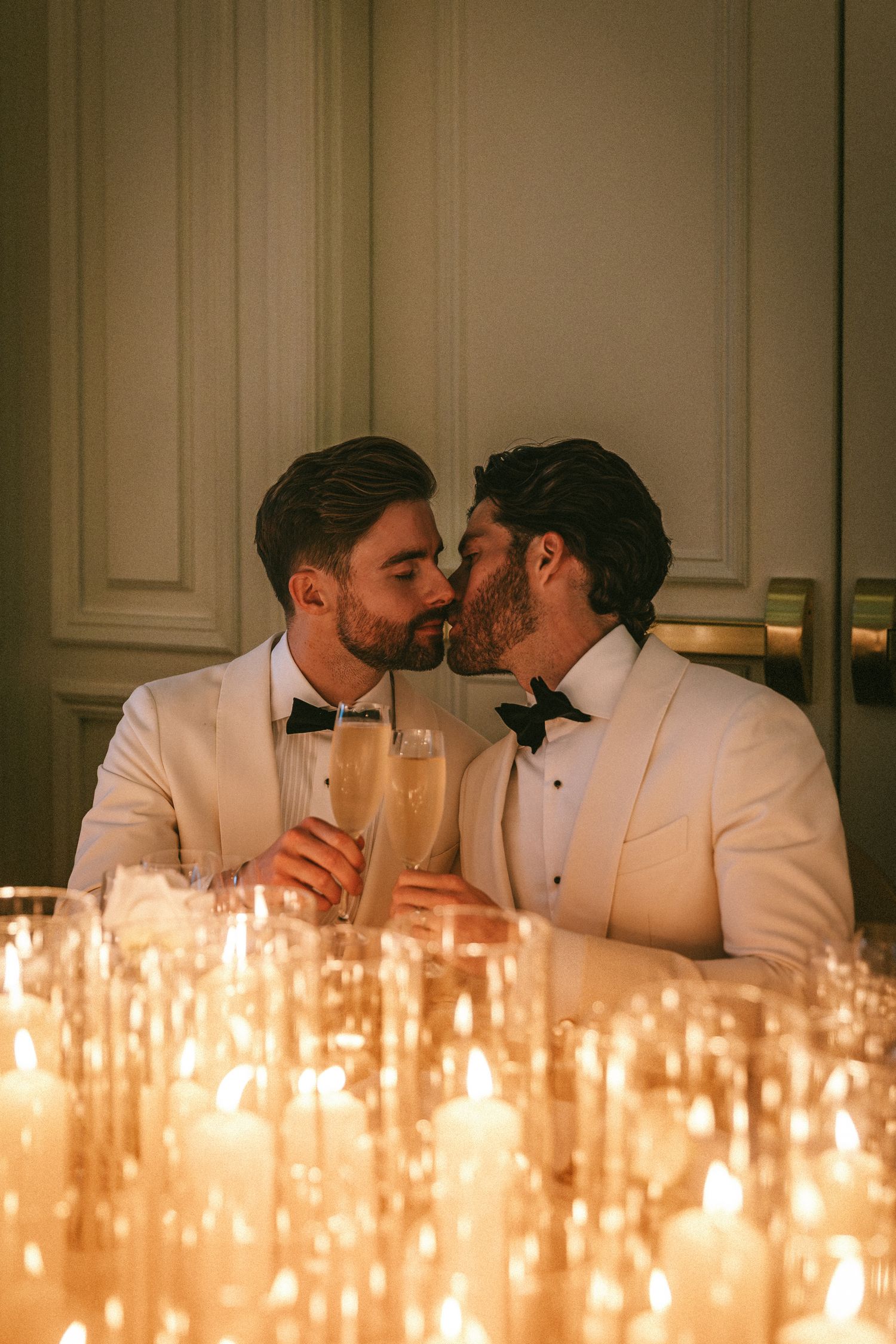 Two grooms in white tuxedos toast champagne surrounded by glowing candles at wedding reception.