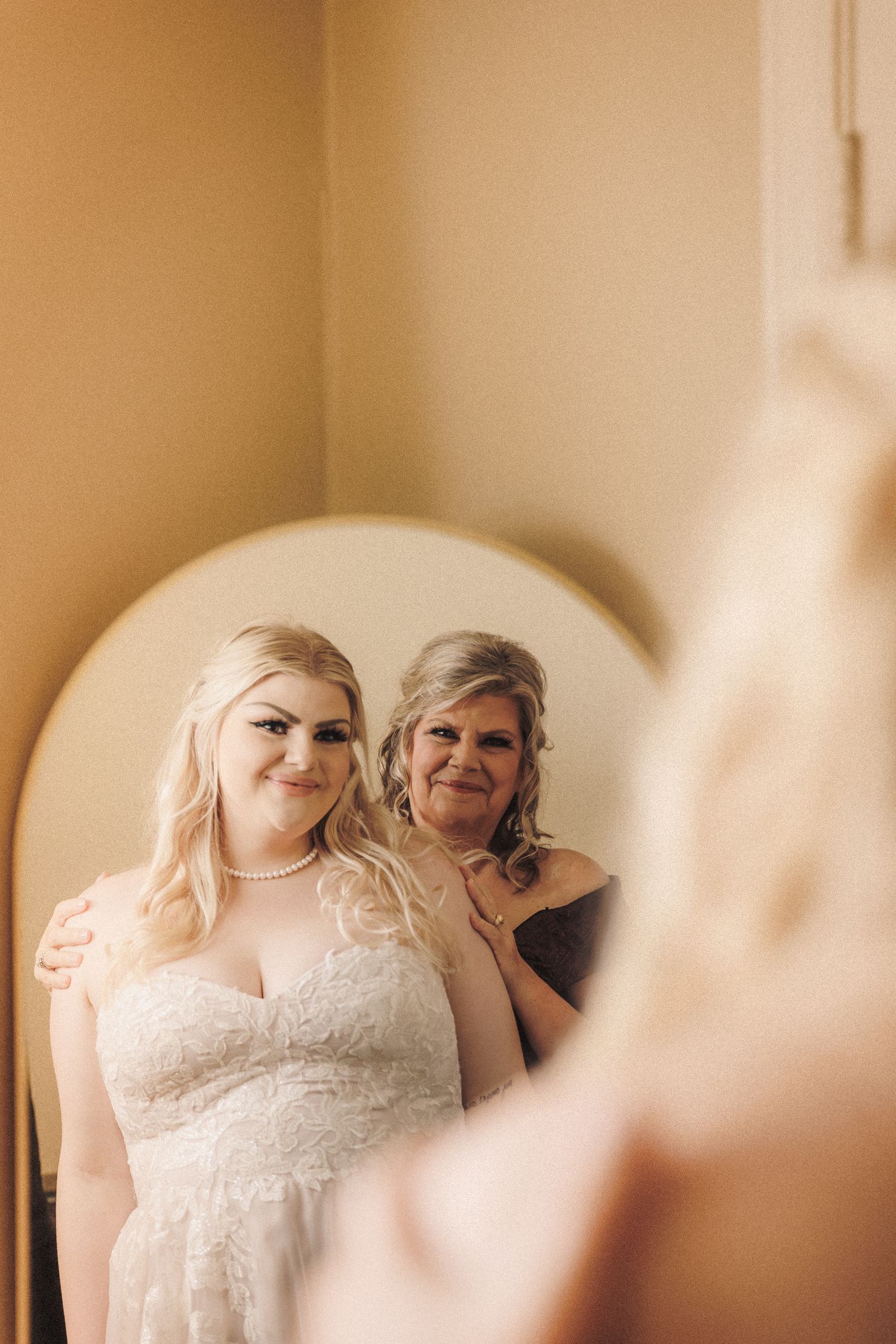 Bride in lace wedding gown and mother sharing a tender mirror reflection moment together.