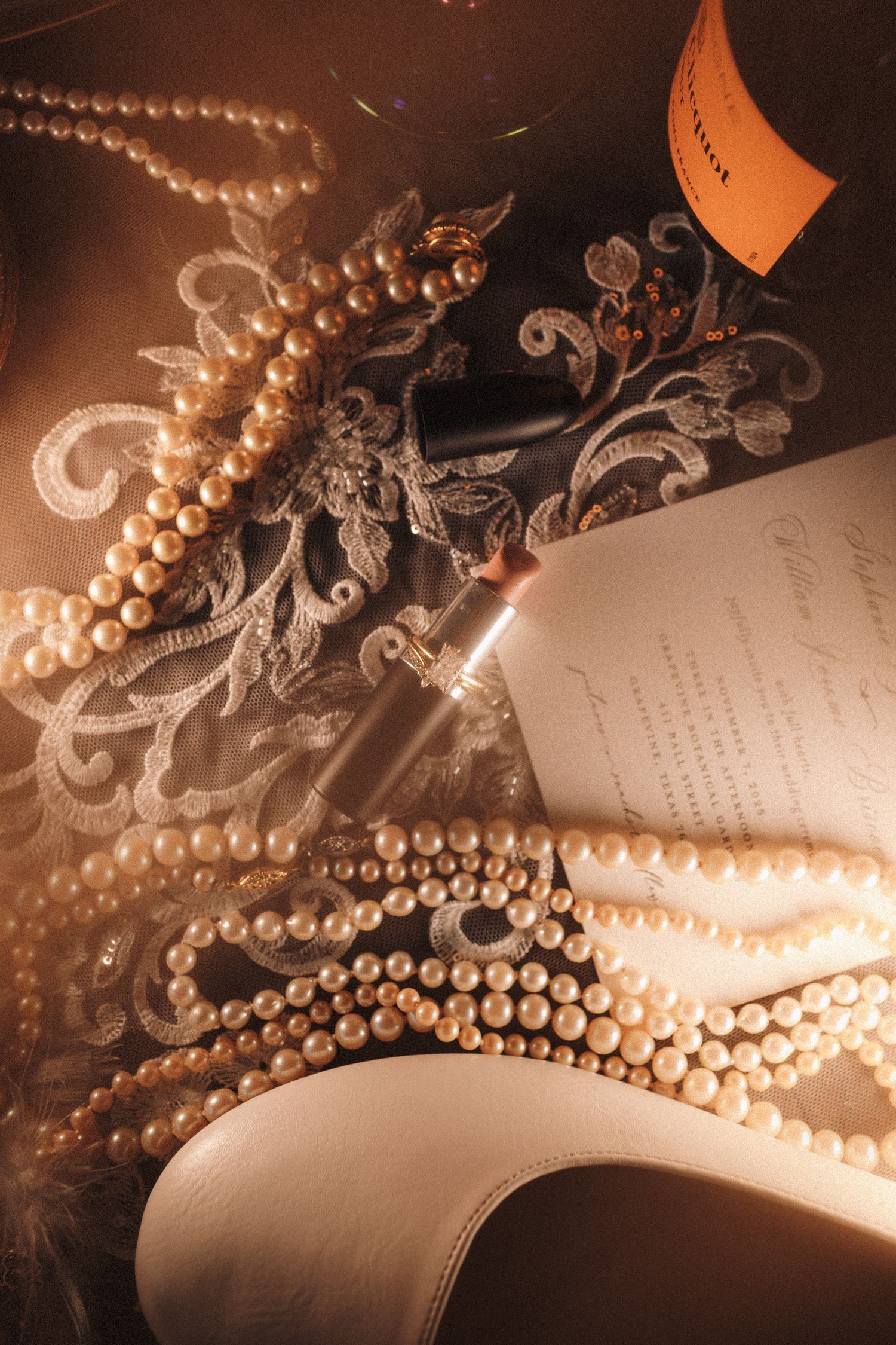 Elegant flat lay of pearl necklaces, champagne bottle, lace fabric, lipstick, and wedding invitation in warm golden light.