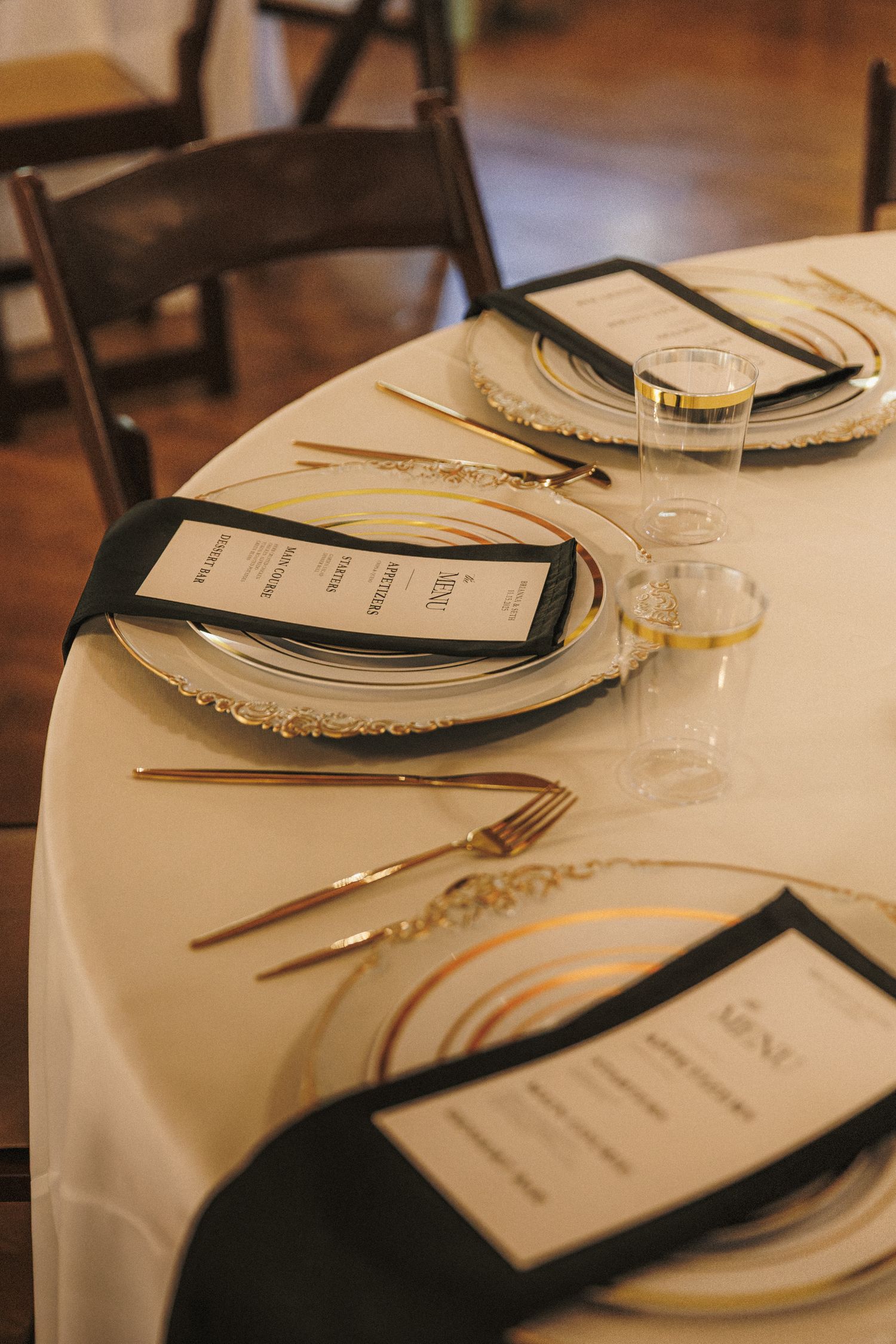 Elegant wedding reception table setting with gold cutlery, charger plates, and black menu cards on white linen.