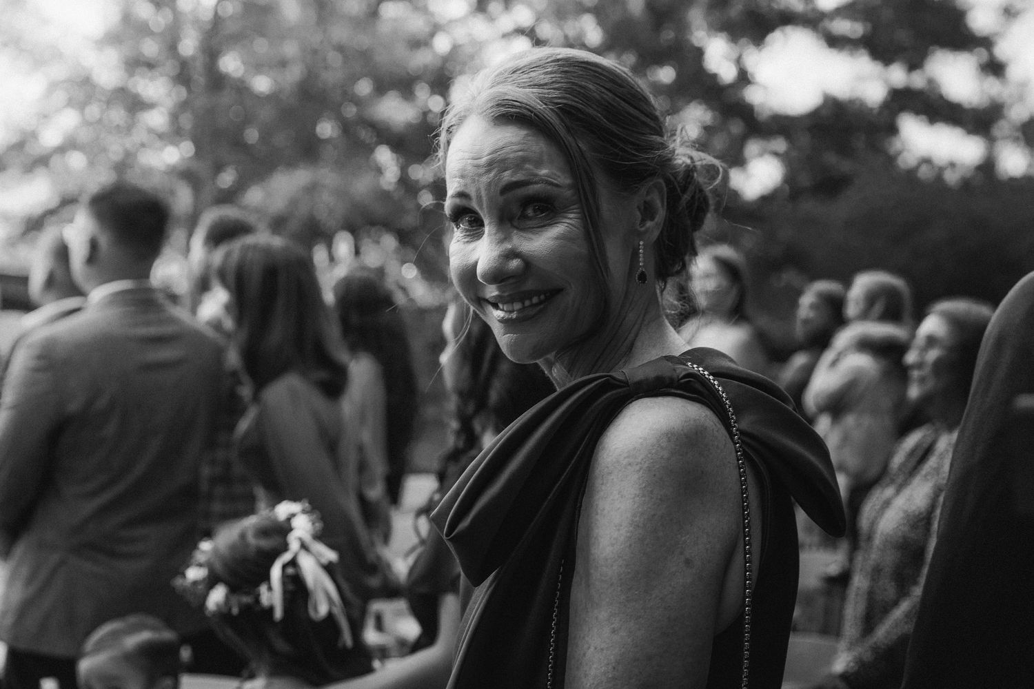 Black and white portrait of smiling woman at outdoor wedding reception with guests in background.