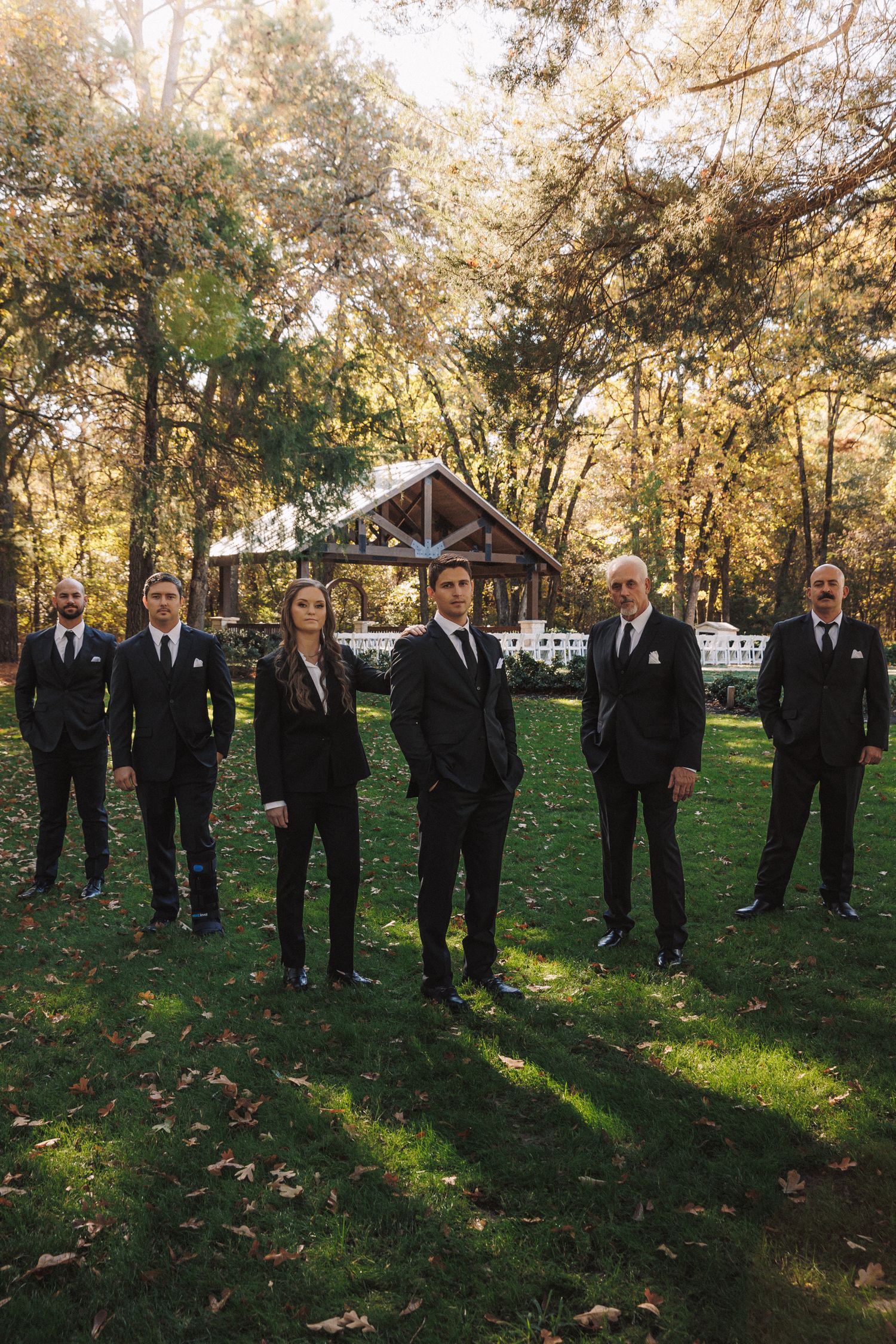 Groom and groomsmen in matching black suits posing outdoors with autumn trees and pavilion backdrop.