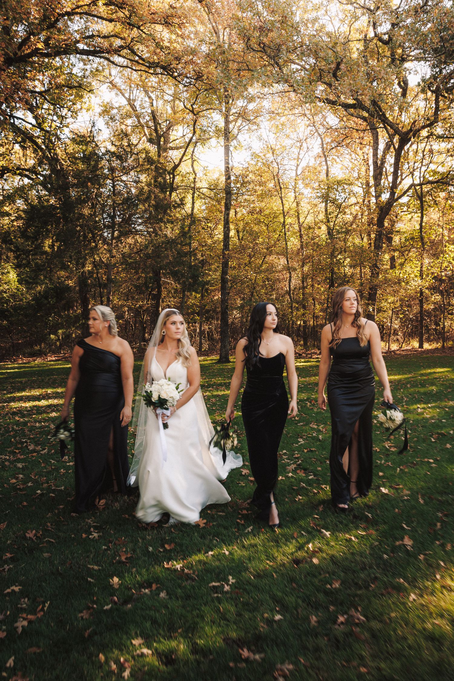 Bride in white gown walks with three bridesmaids in black dresses carrying bouquets in a sunlit forest.