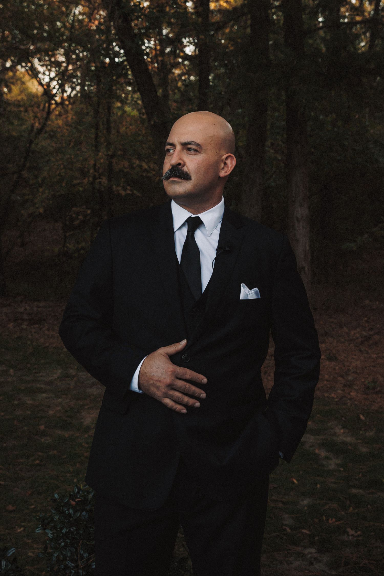 Bald man with mustache in black suit and tie posing outdoors in a wooded setting.