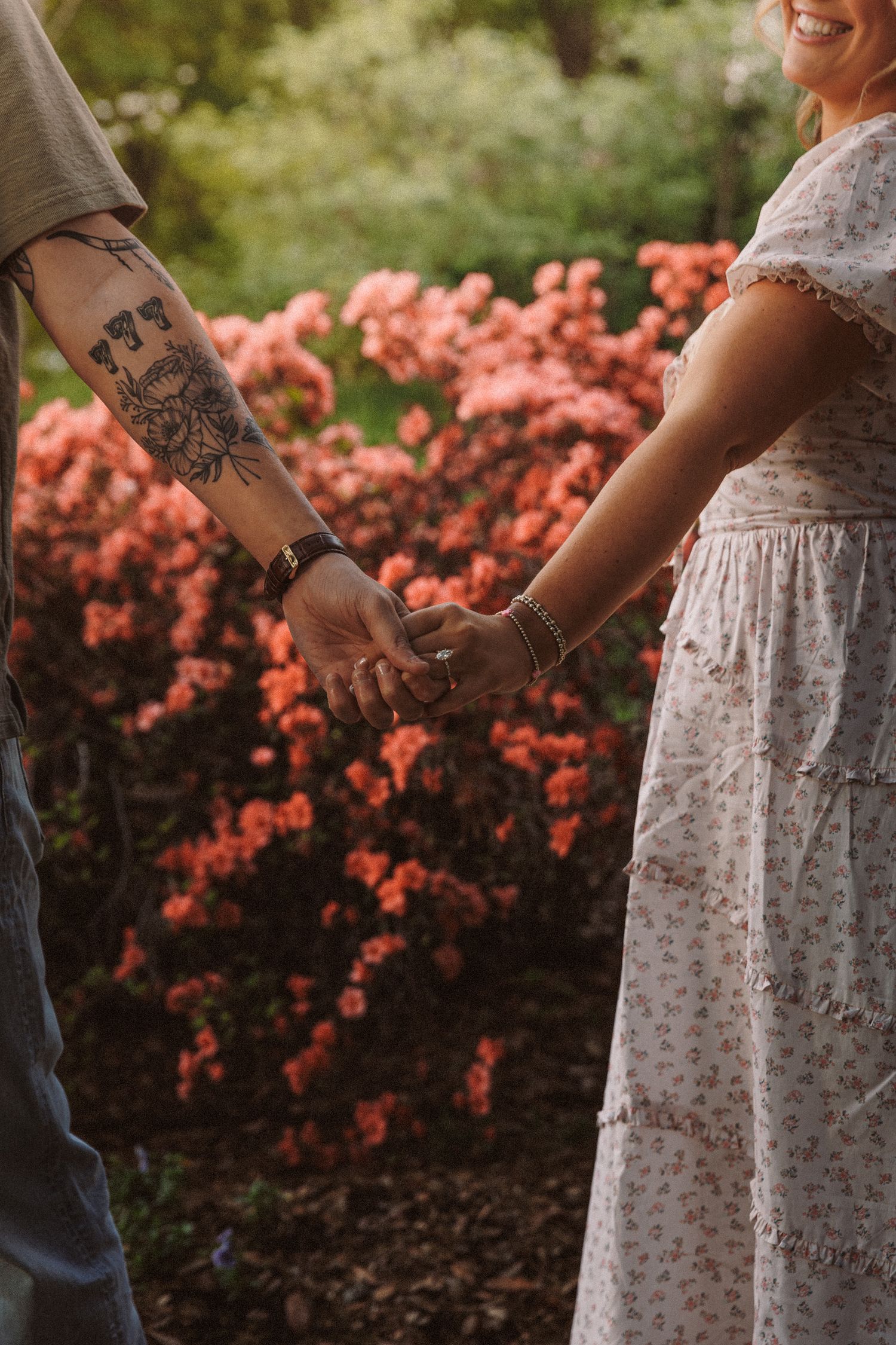 Couple holding hands in a garden with pink flowering bushes, man with tattoed arm wearing a watch.