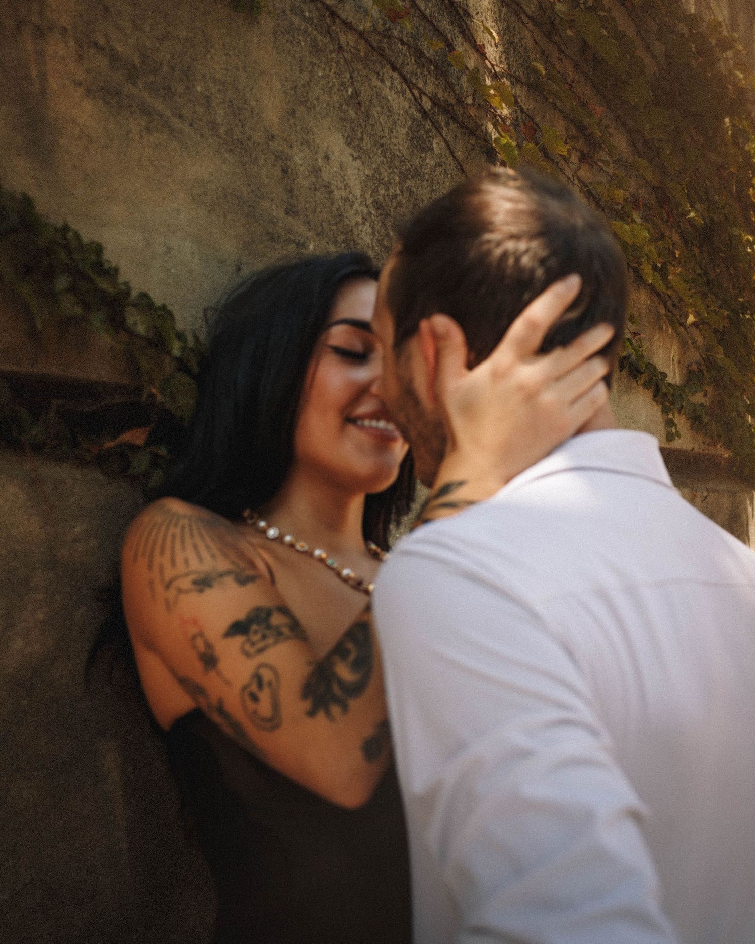 A romantic couple embraces against a textured wall, with the person in black wearing a pearl necklace and visible tattoos.
