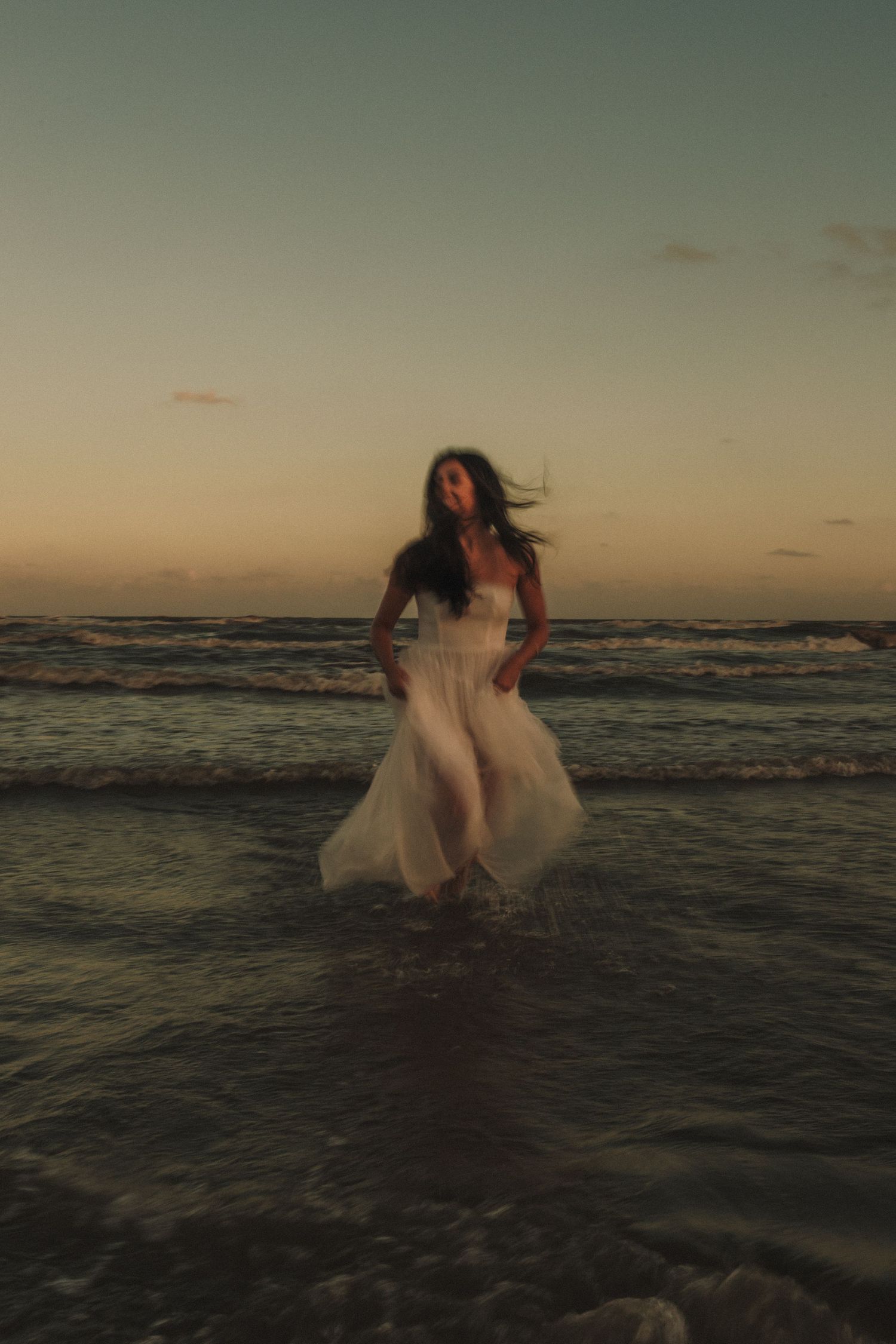 A couple in wedding attire embraces in the ocean waves during sunset creating a romantic beach scene.