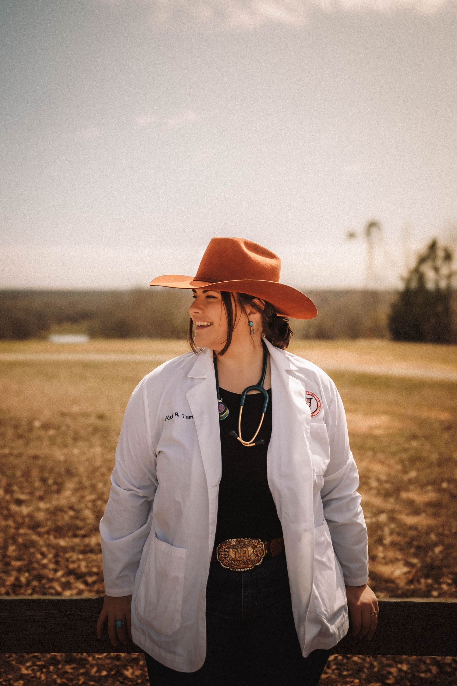 A person in a white medical coat and orange cowboy hat stands in a field at sunset looking contemplative.