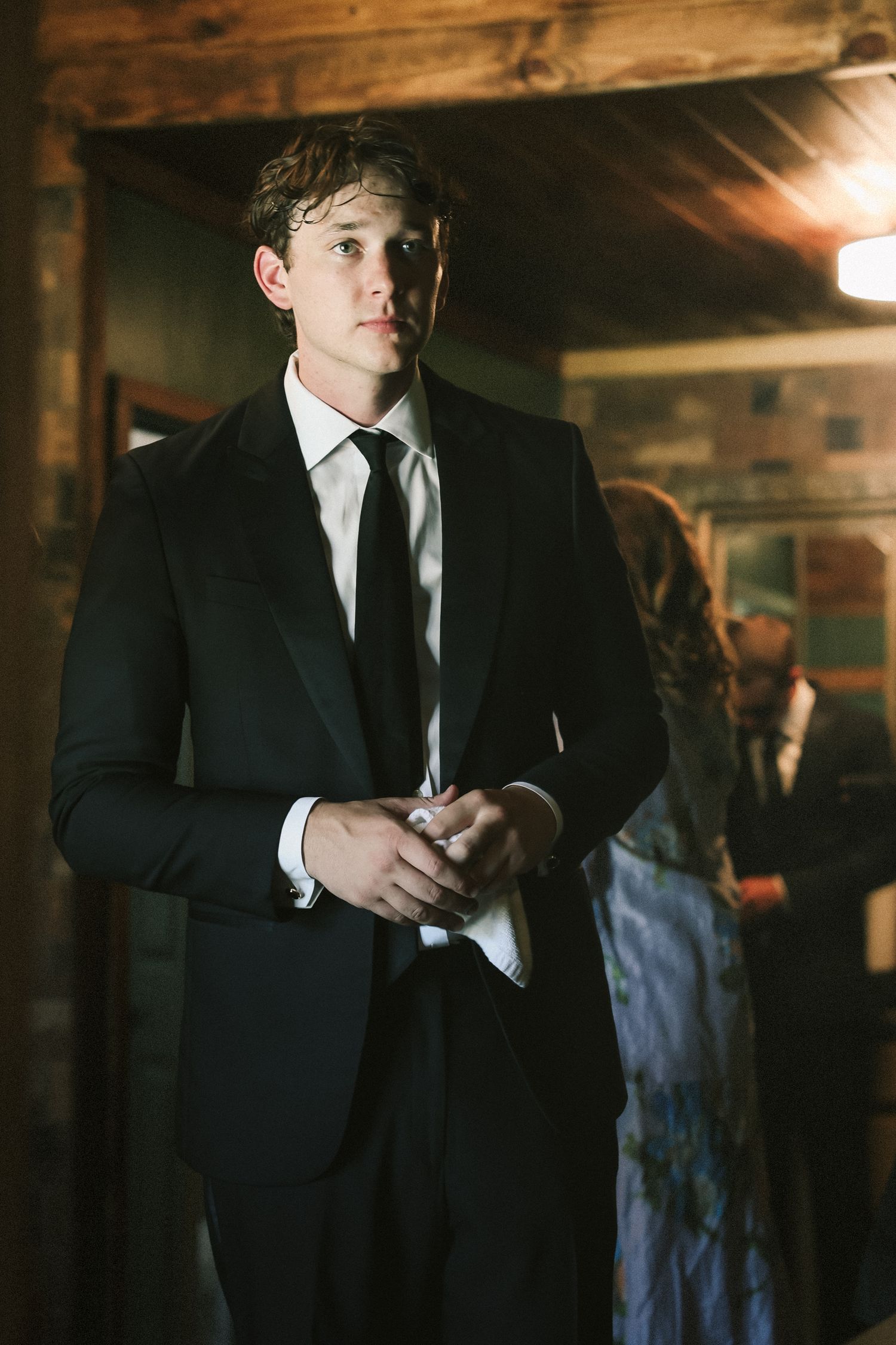 Man wearing black suit and tie adjusts his sleeve in dimly lit room.