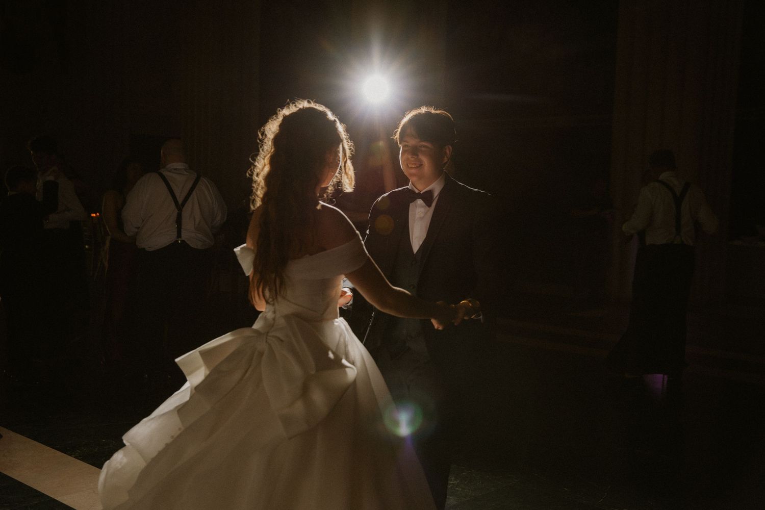 Bride and groom sharing first dance in spotlight at elegant dark wedding reception.