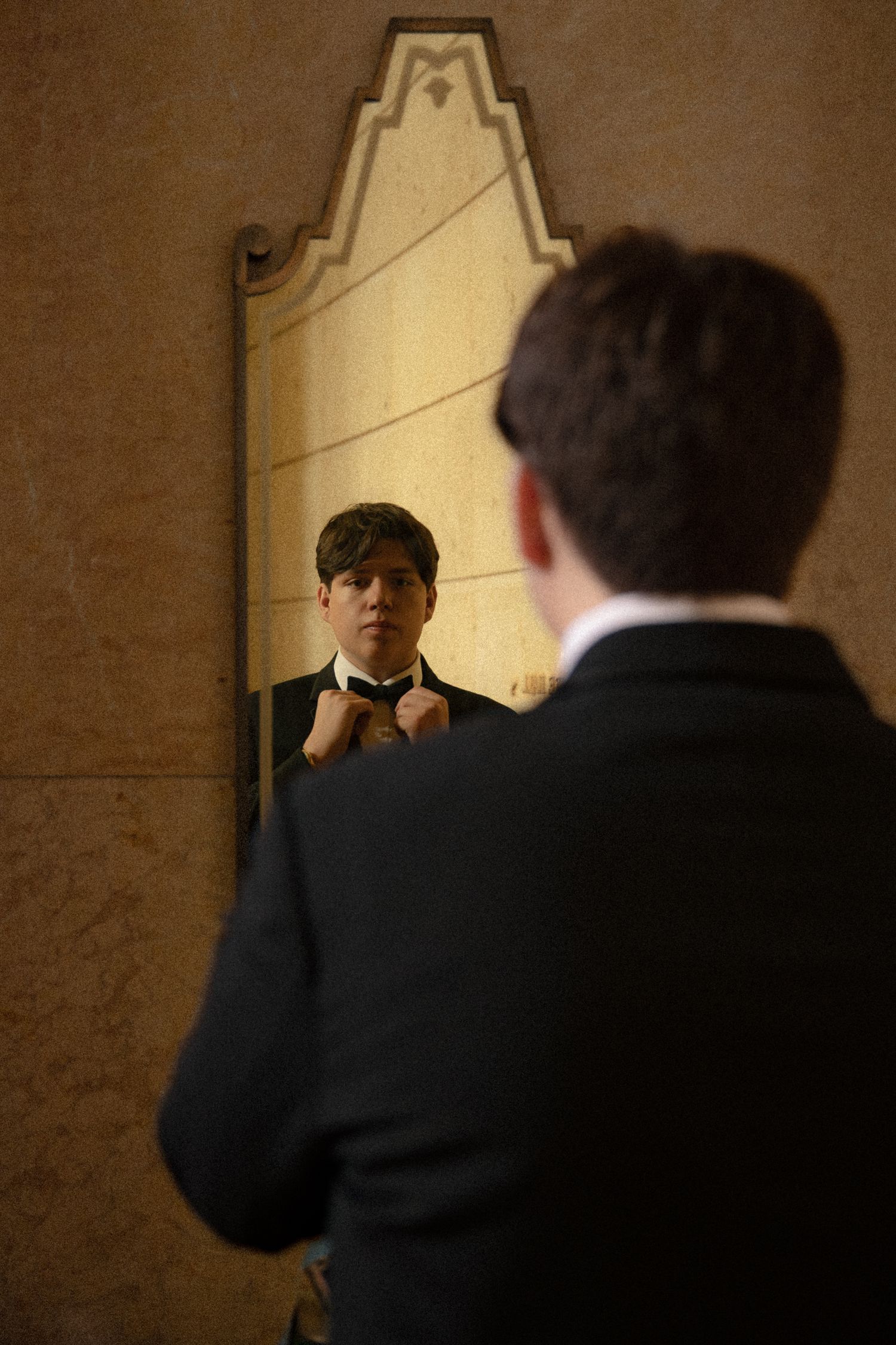 Groom in black tuxedo adjusting bow tie while looking at reflection in ornate art deco mirror.