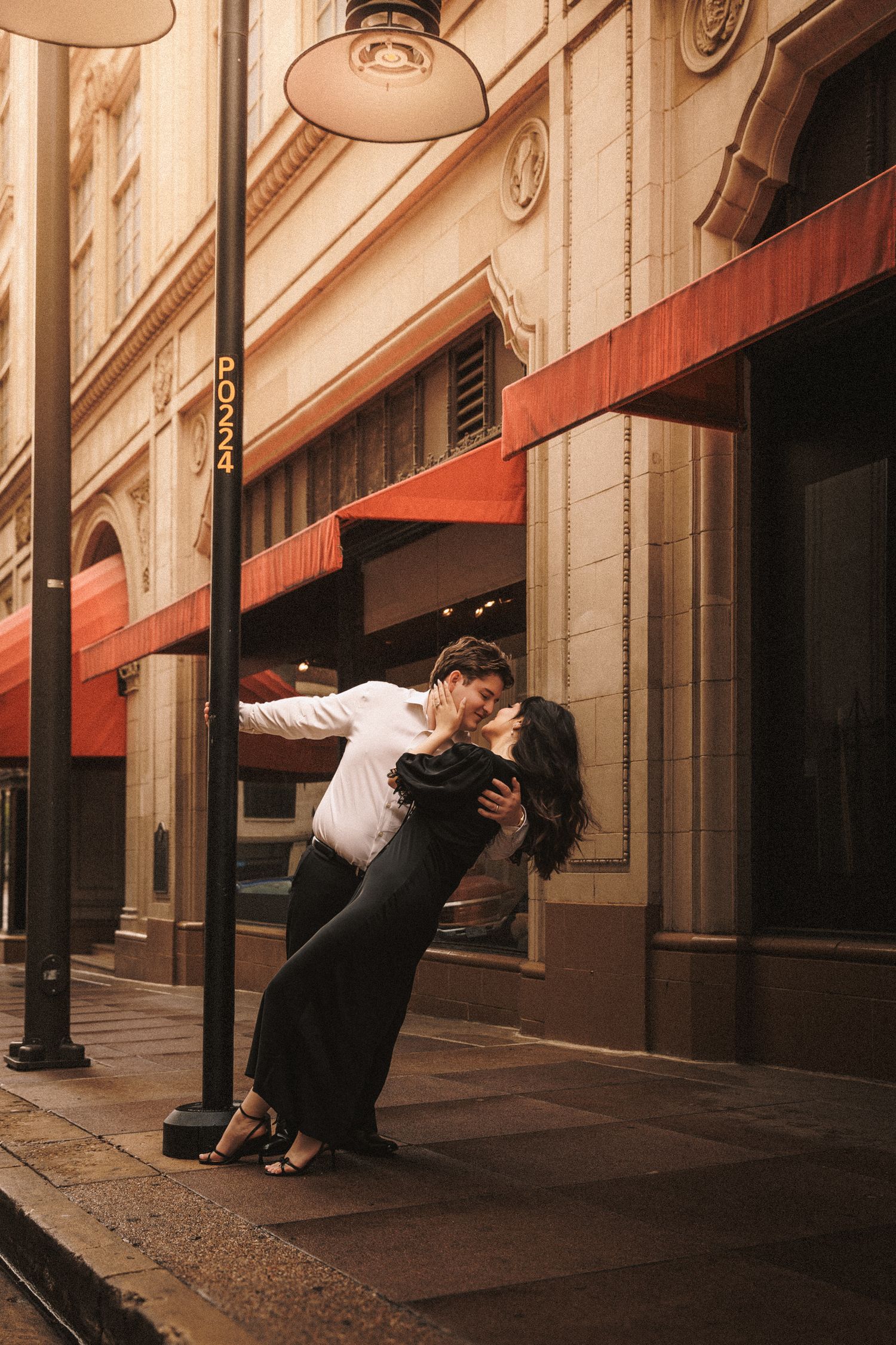 Romantic couple sharing a dip kiss beside a street pole outside a building with red awnings at dusk.