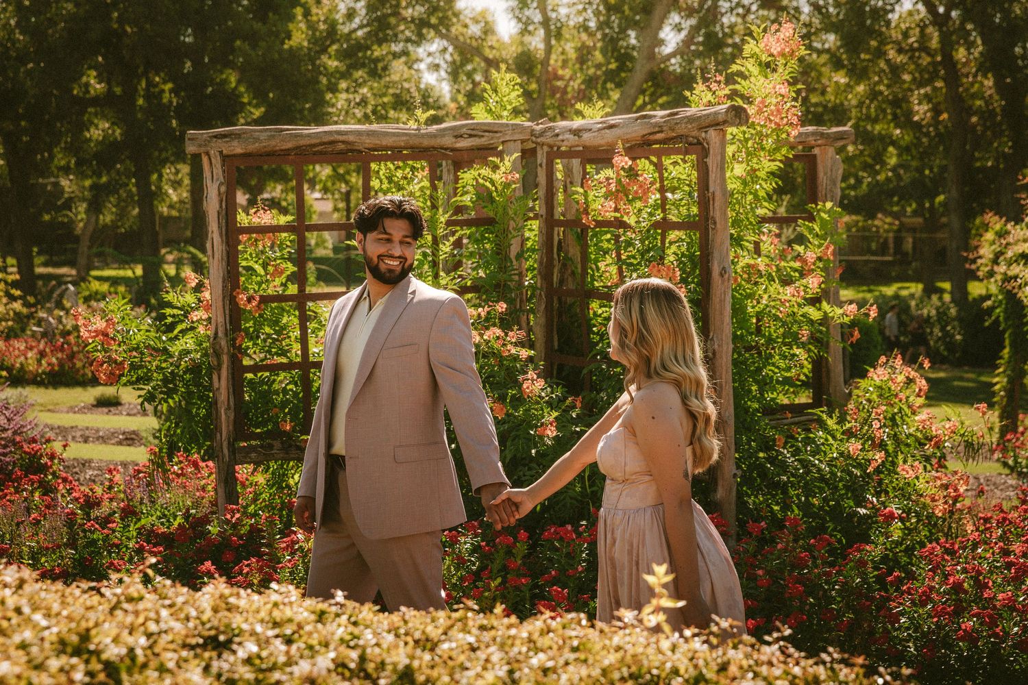 A couple strolls through a lush garden with blooming flowers during sunset.