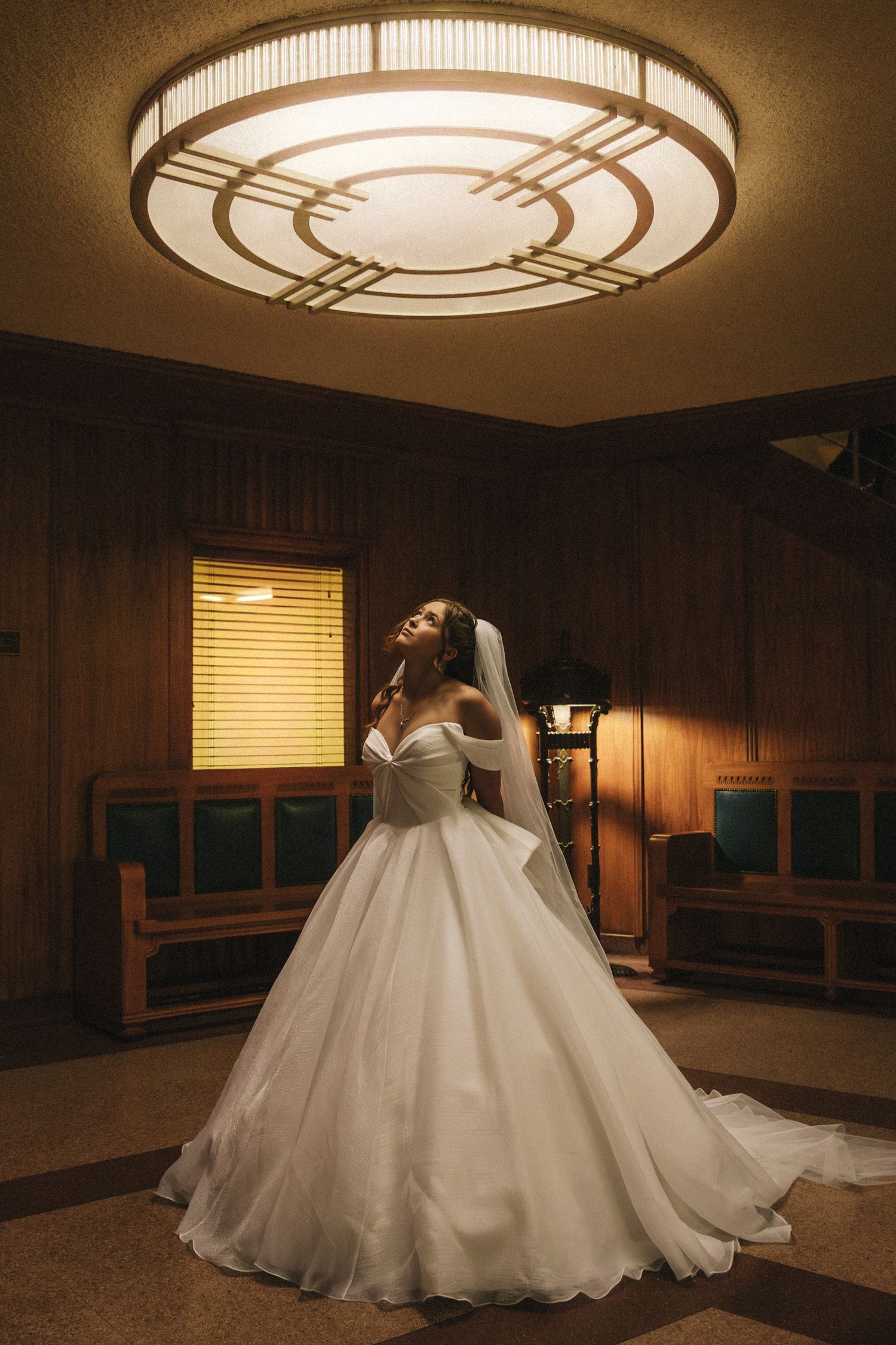 Bride in strapless ball gown wedding dress posing under ornate ceiling light in wood-paneled room.