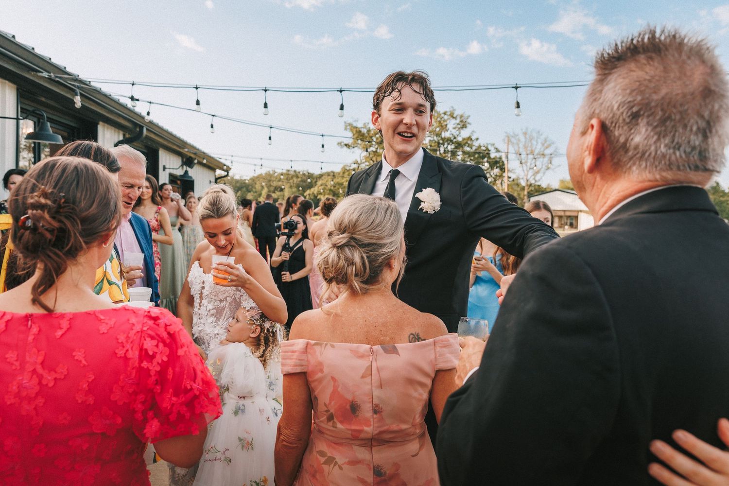 Wedding guests laugh and celebrate during an outdoor reception under string lights.