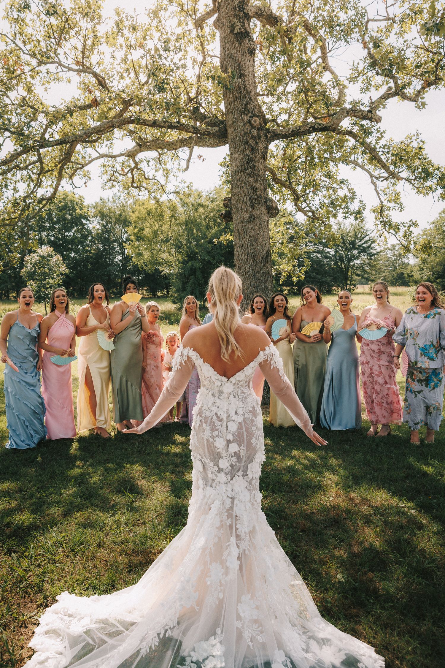 A wedding party wearing pastel-colored dresses poses in a line under a canopy of trees during an outdoor celebration.