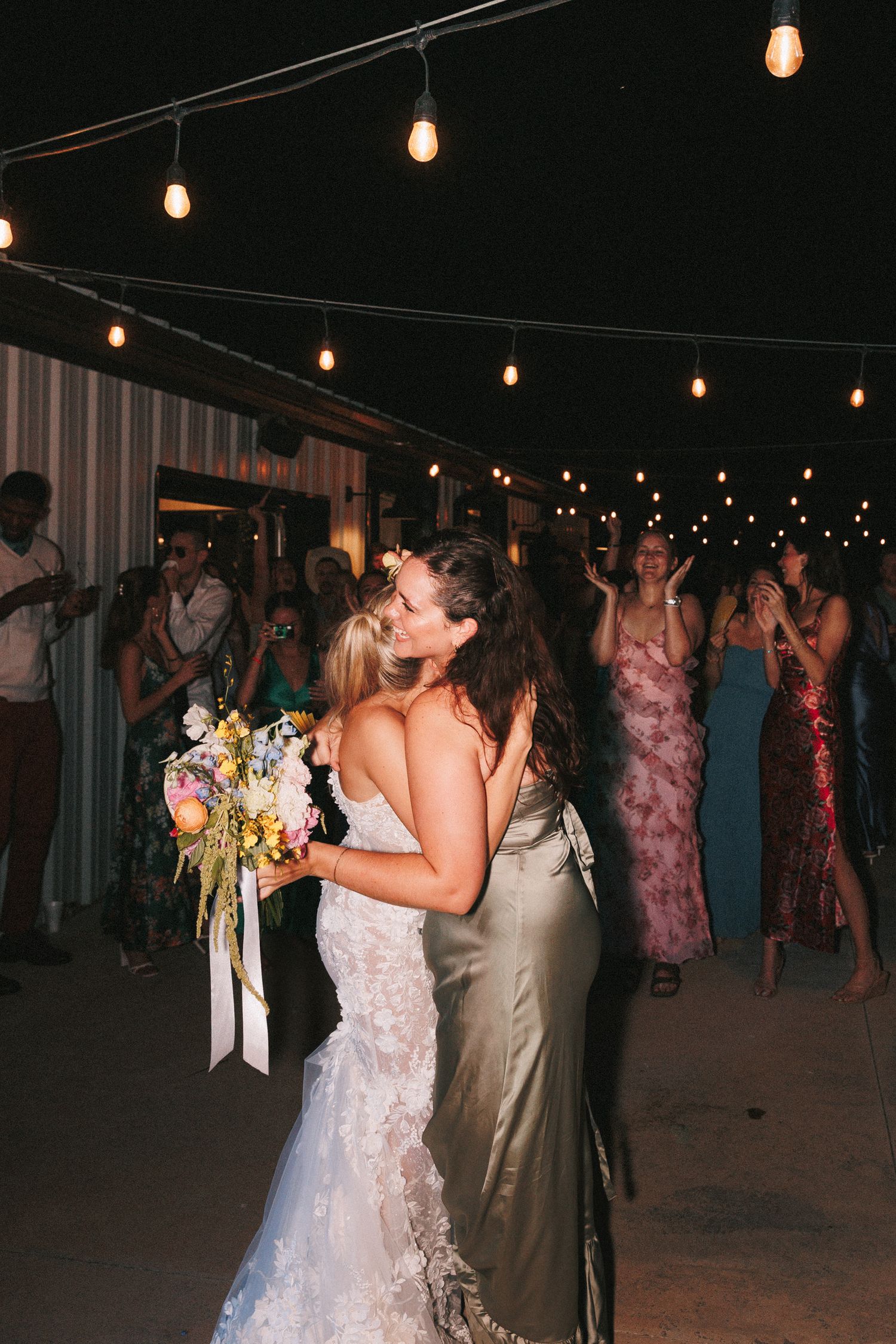 Wedding guests dance under string lights at an outdoor evening reception.