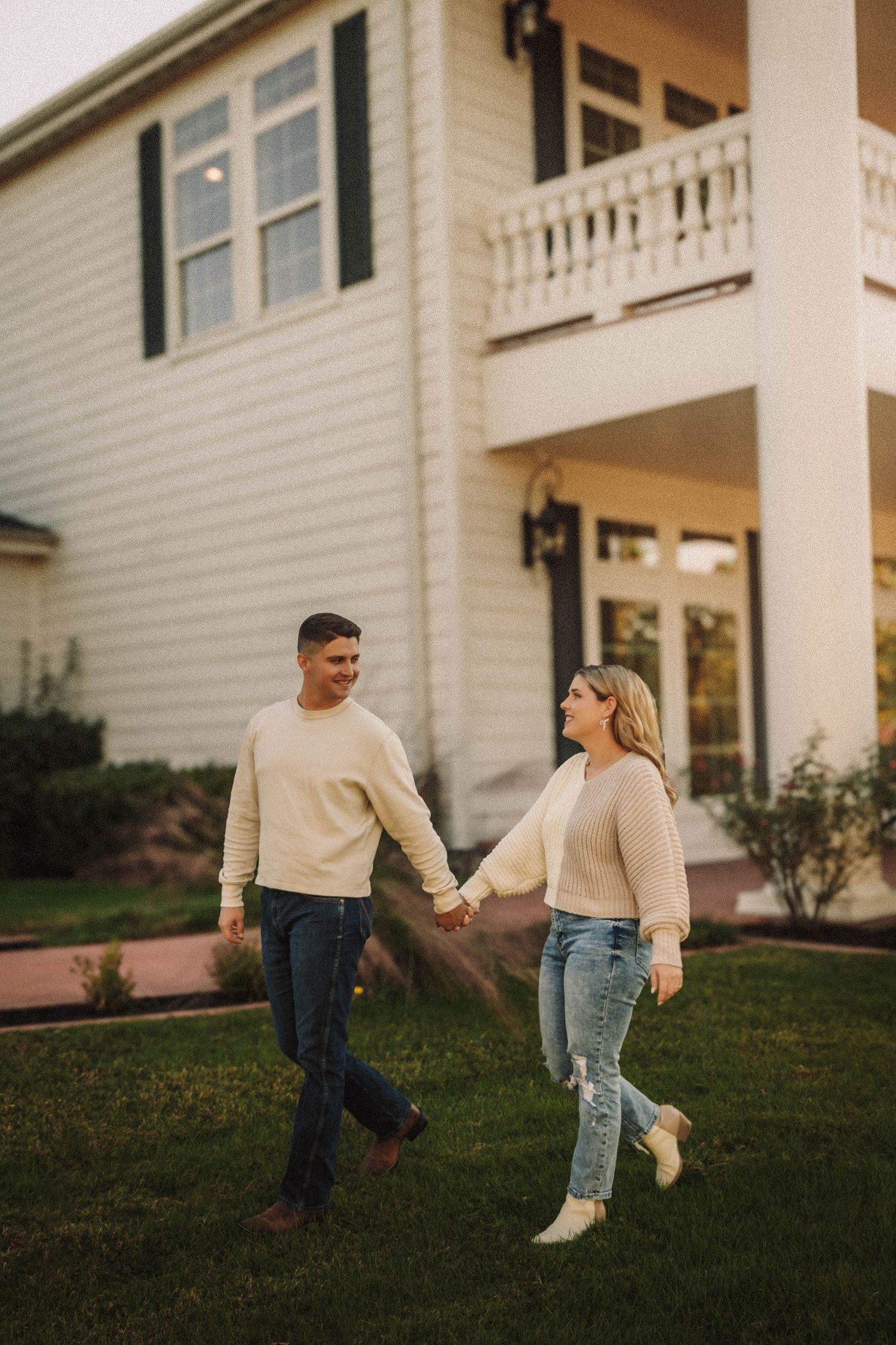 A couple in matching cream sweaters dance playfully on the lawn of a white house with a balcony at sunset.