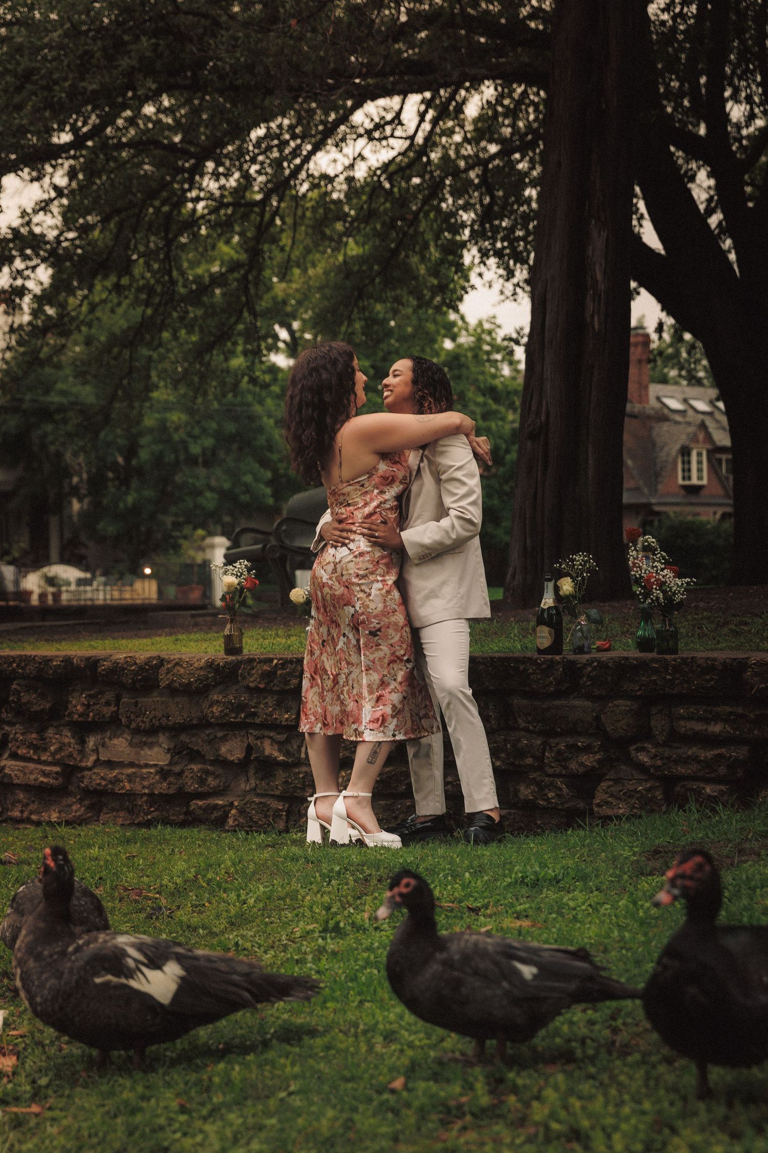 A couple poses near a stone wall while black ducks graze on the grass in a park setting.