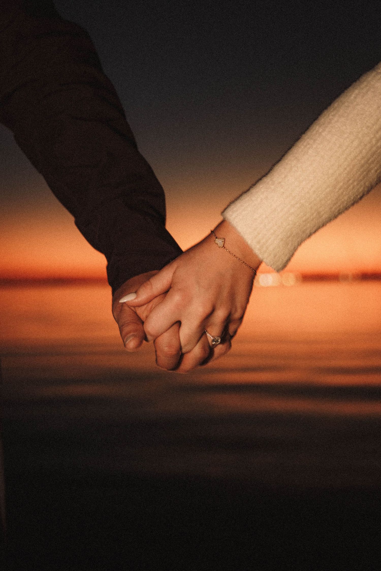 A close-up shot of two people holding hands at sunset, with one wearing an engagement ring.