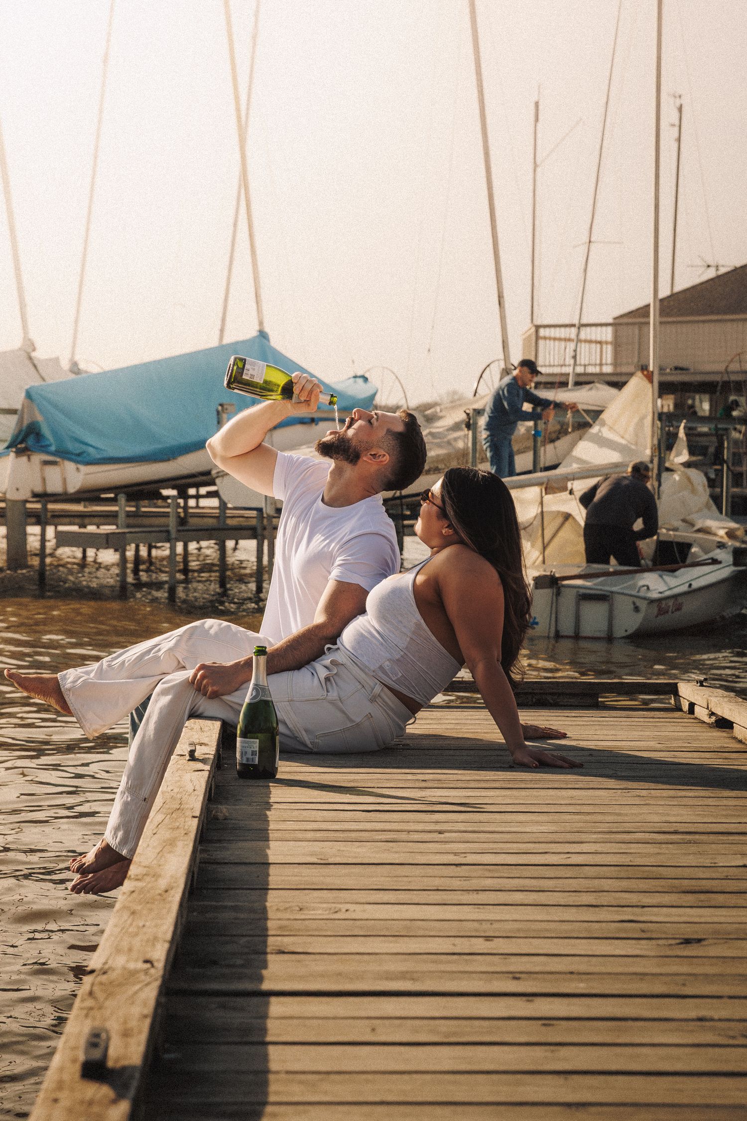 Couple relaxing and drinking wine together on a wooden dock by a marina with sailboats at sunset.