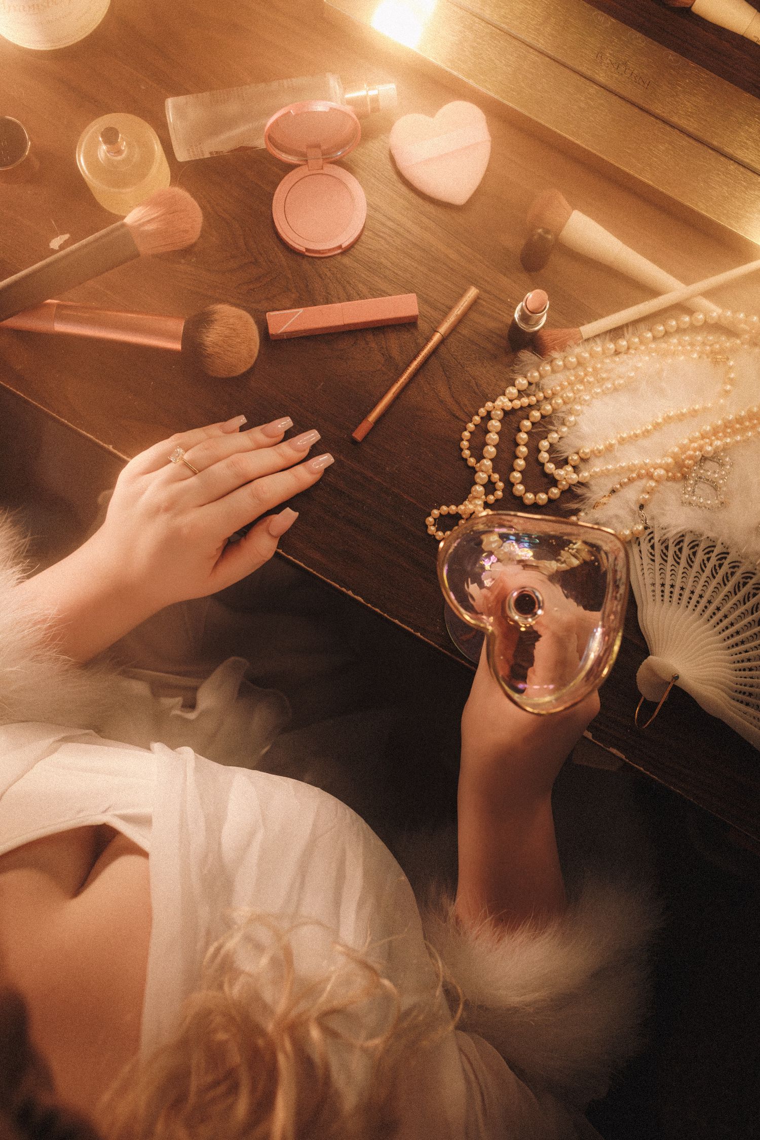 Bride at vanity mirror with makeup, pearls, and perfume in warm glowing light wearing white robe.