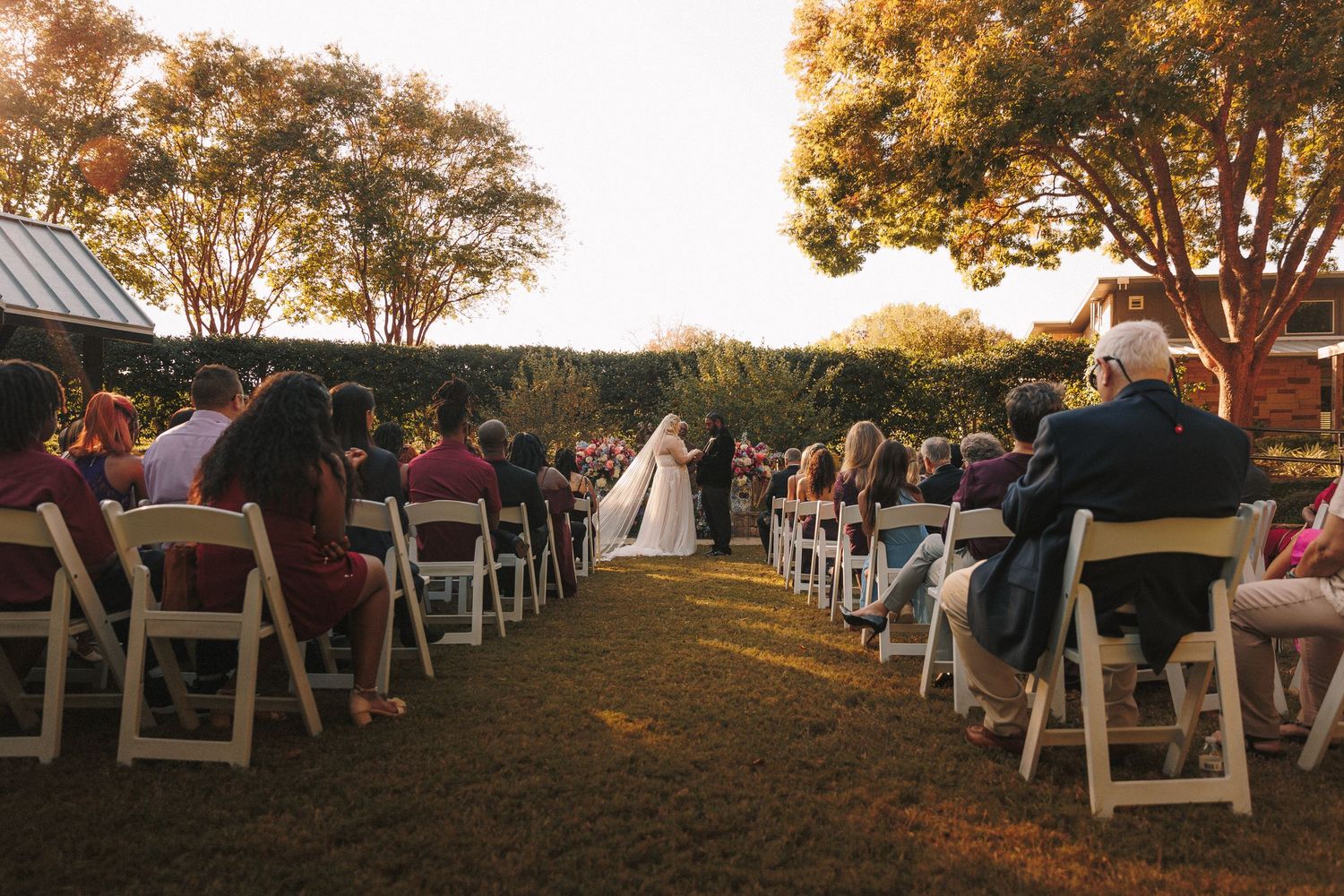 Outdoor wedding ceremony at sunset with guests seated in white chairs along a garden aisle.