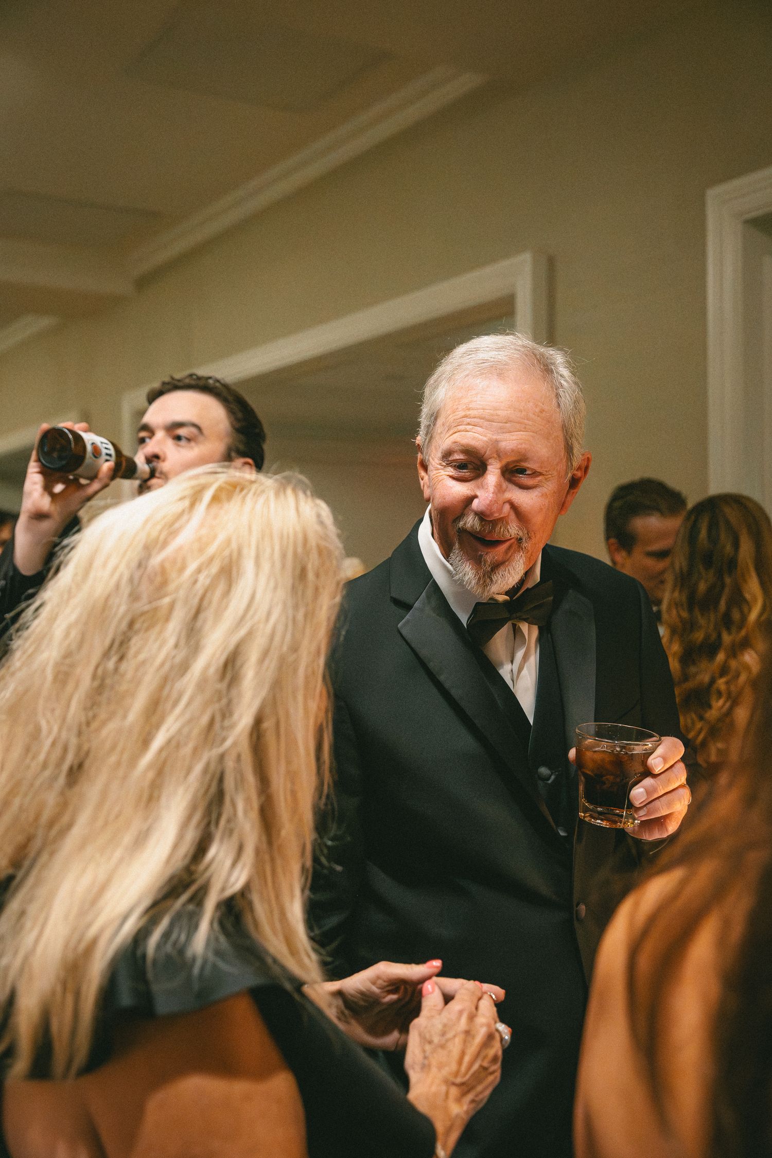 Elderly wedding guest in black tuxedo smiles while holding a drink at a wedding reception.
