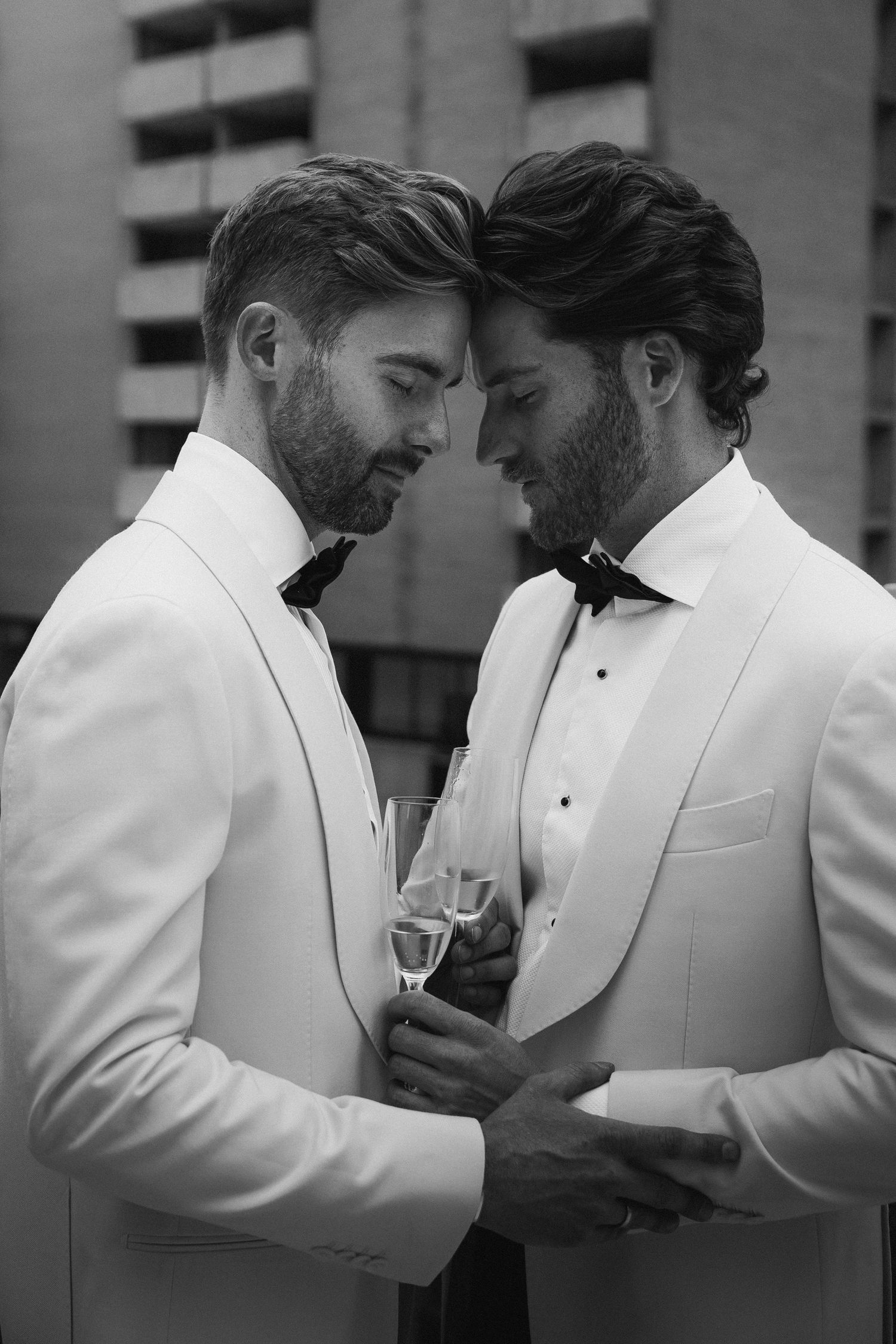 Two grooms in white tuxedos with bow ties sharing champagne in an intimate black and white portrait.
