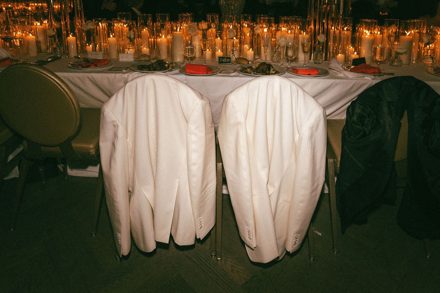 Two white tuxedo jackets draped over chairs at a candlelit wedding reception dinner table.