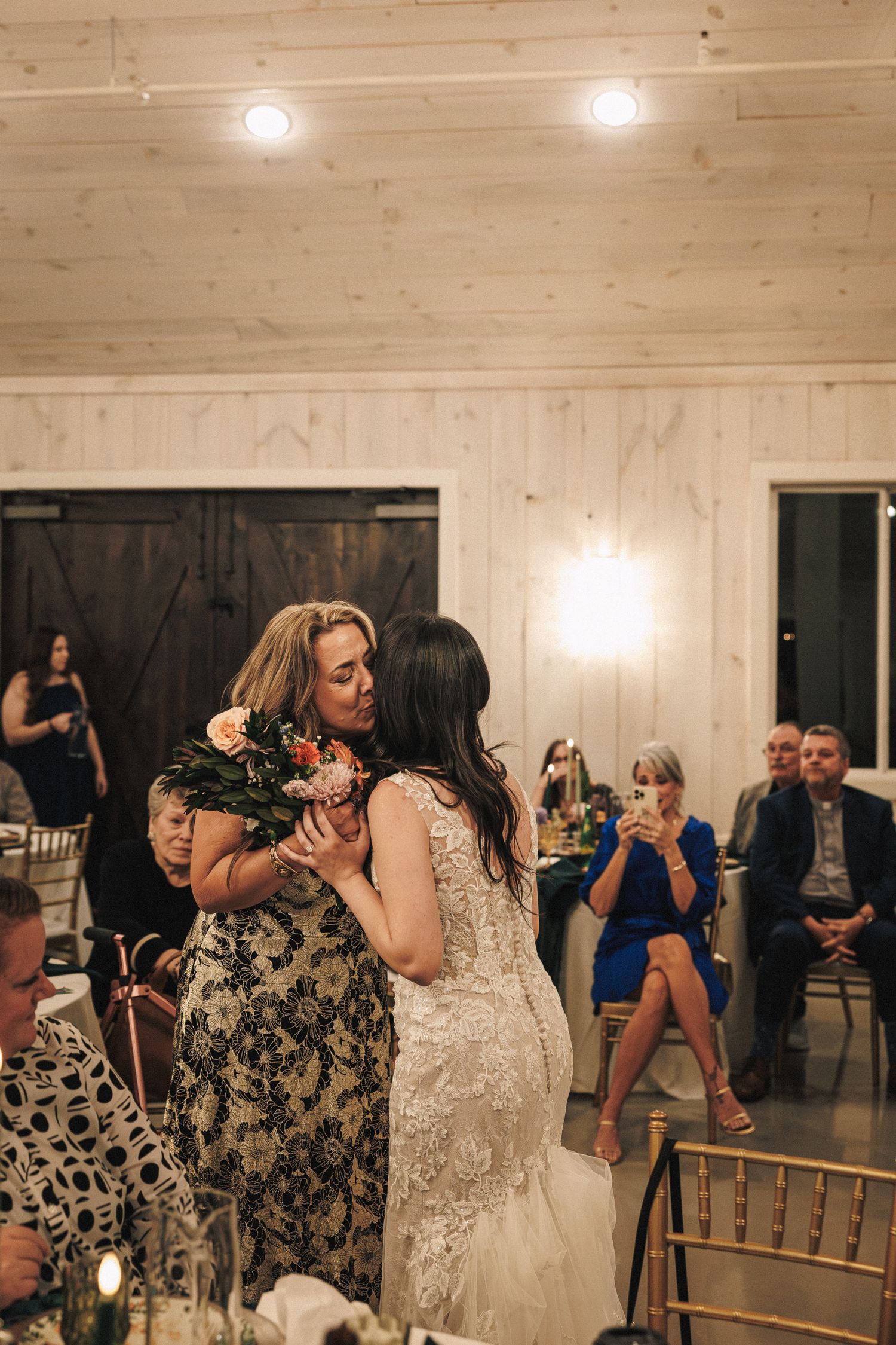 Bride in lace gown shares an emotional hug with her mother during the wedding reception in a barn venue.