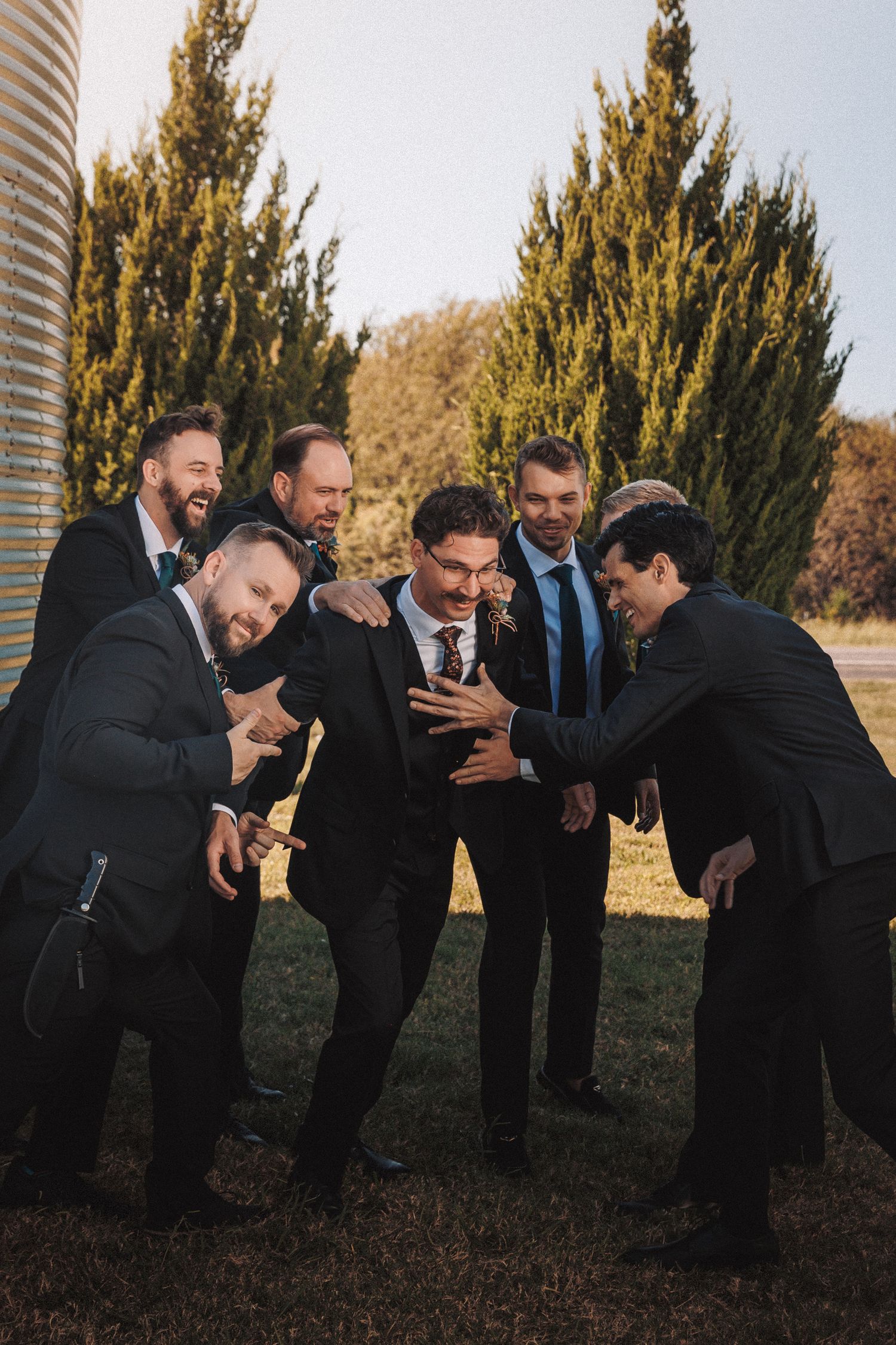 Groom surrounded by groomsmen in black suits sharing a joyful moment outdoors near evergreen trees.