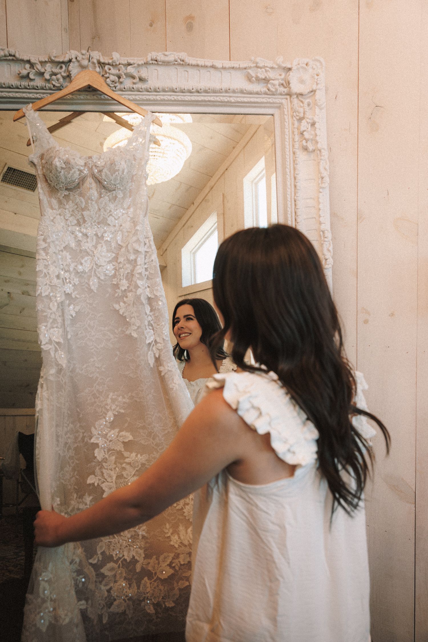 Bride admiring her lace wedding gown hanging on a mirror in a rustic bridal suite.