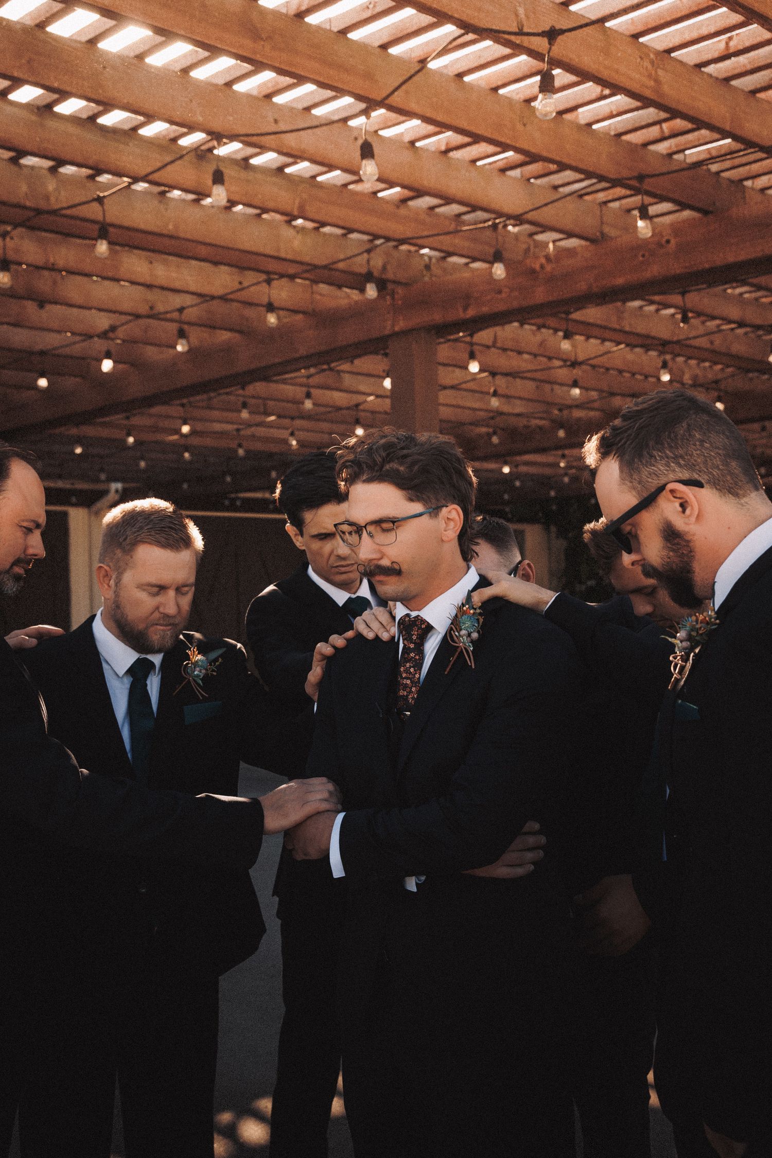 Groomsmen in black suits gather under a pergola with string lights, reading wedding vows together.