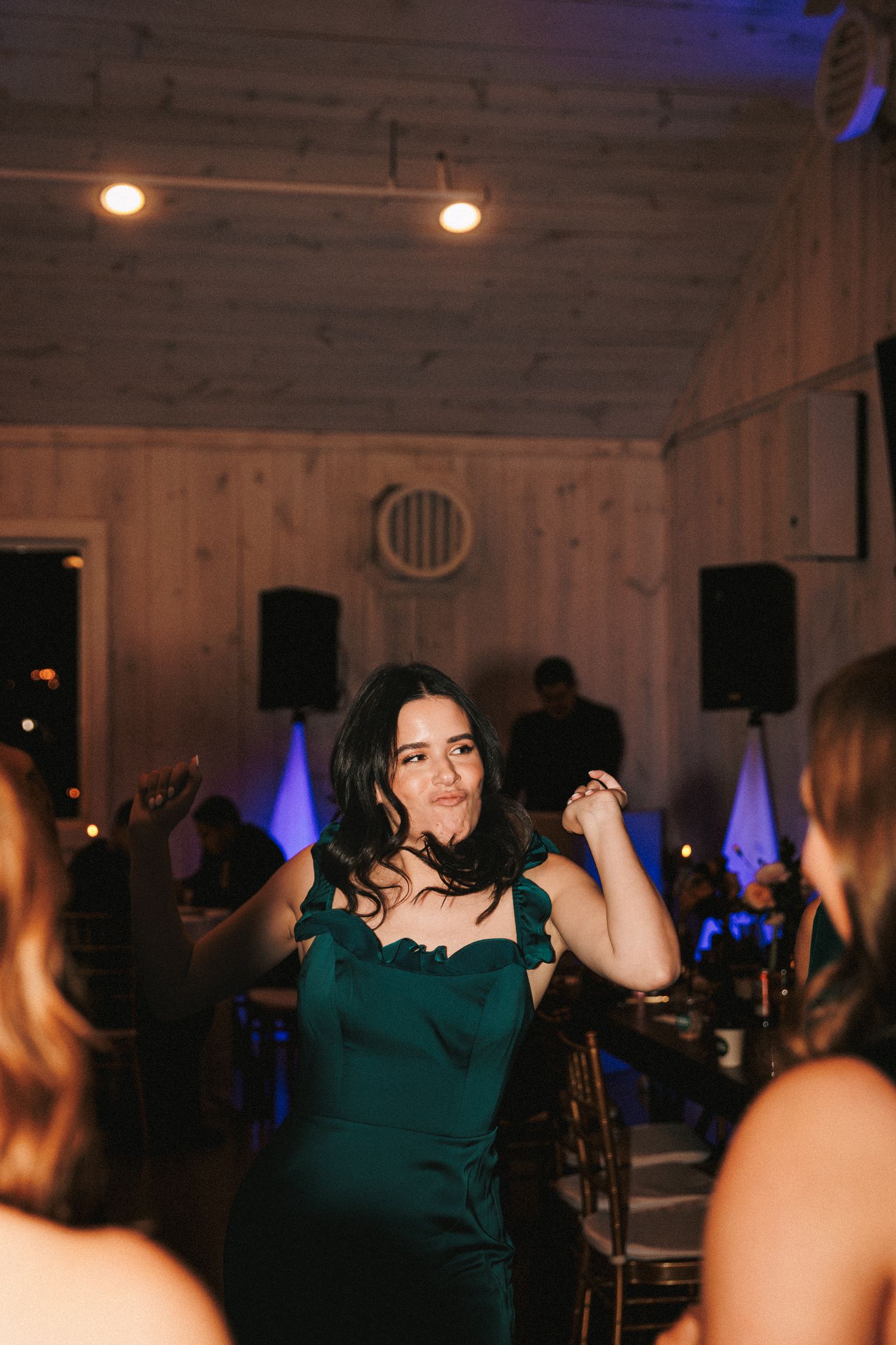 Woman in emerald green dress dances joyfully inside a warmly lit rustic wooden wedding reception hall.